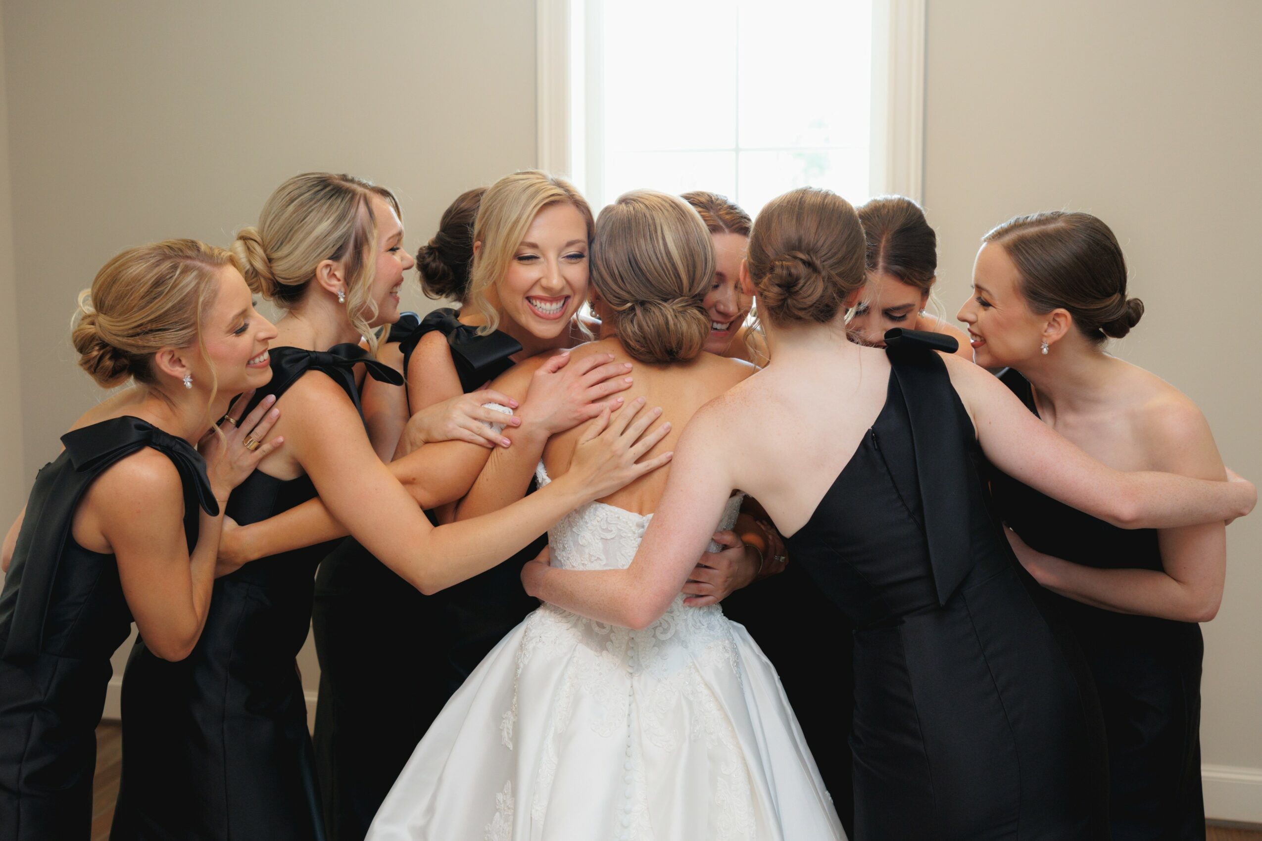 Group of women in black dresses hugging and smiling, with one woman in a white dress, in a bright room.