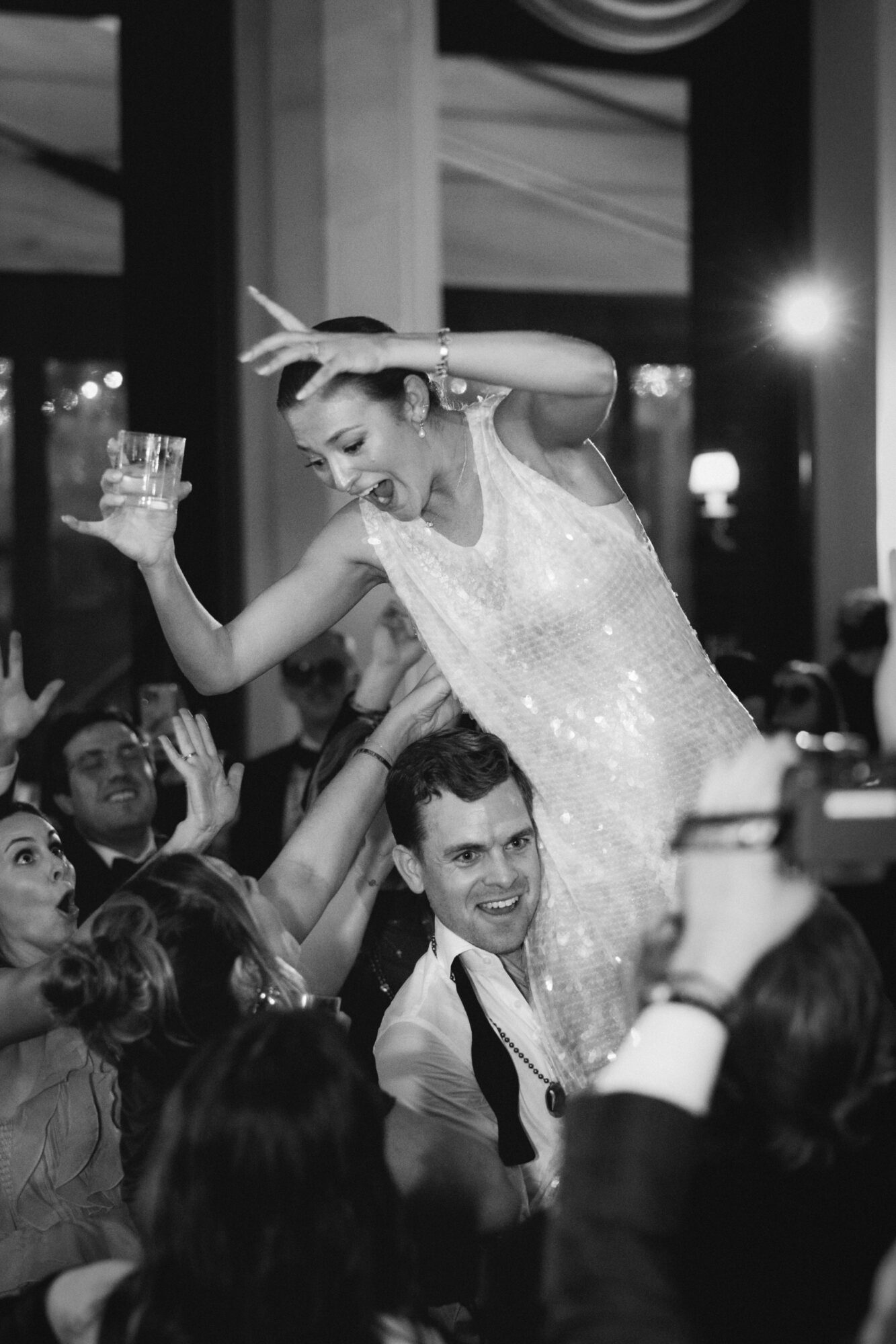 Woman in a dress dancing on a man's shoulders at a celebration, surrounded by people, in a decorated indoor venue.