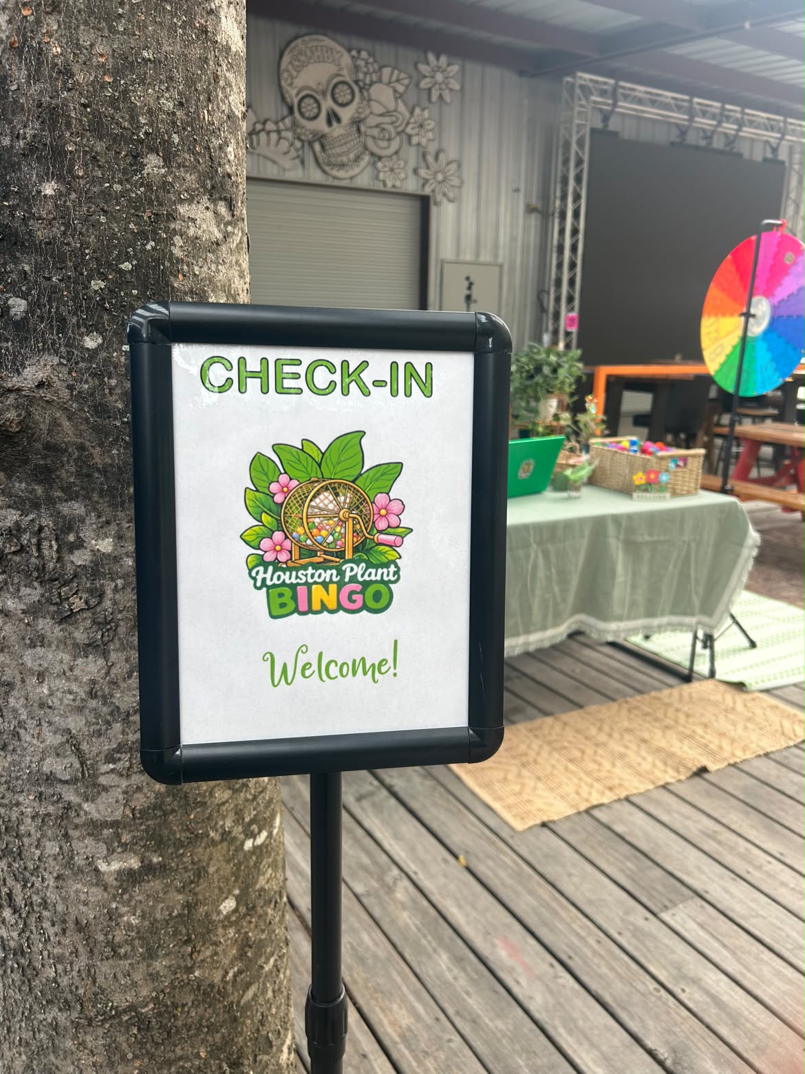 Check-in sign with Houston Plants Bingo logo and welcome message, outdoors with tables and a colorful wheel in background.