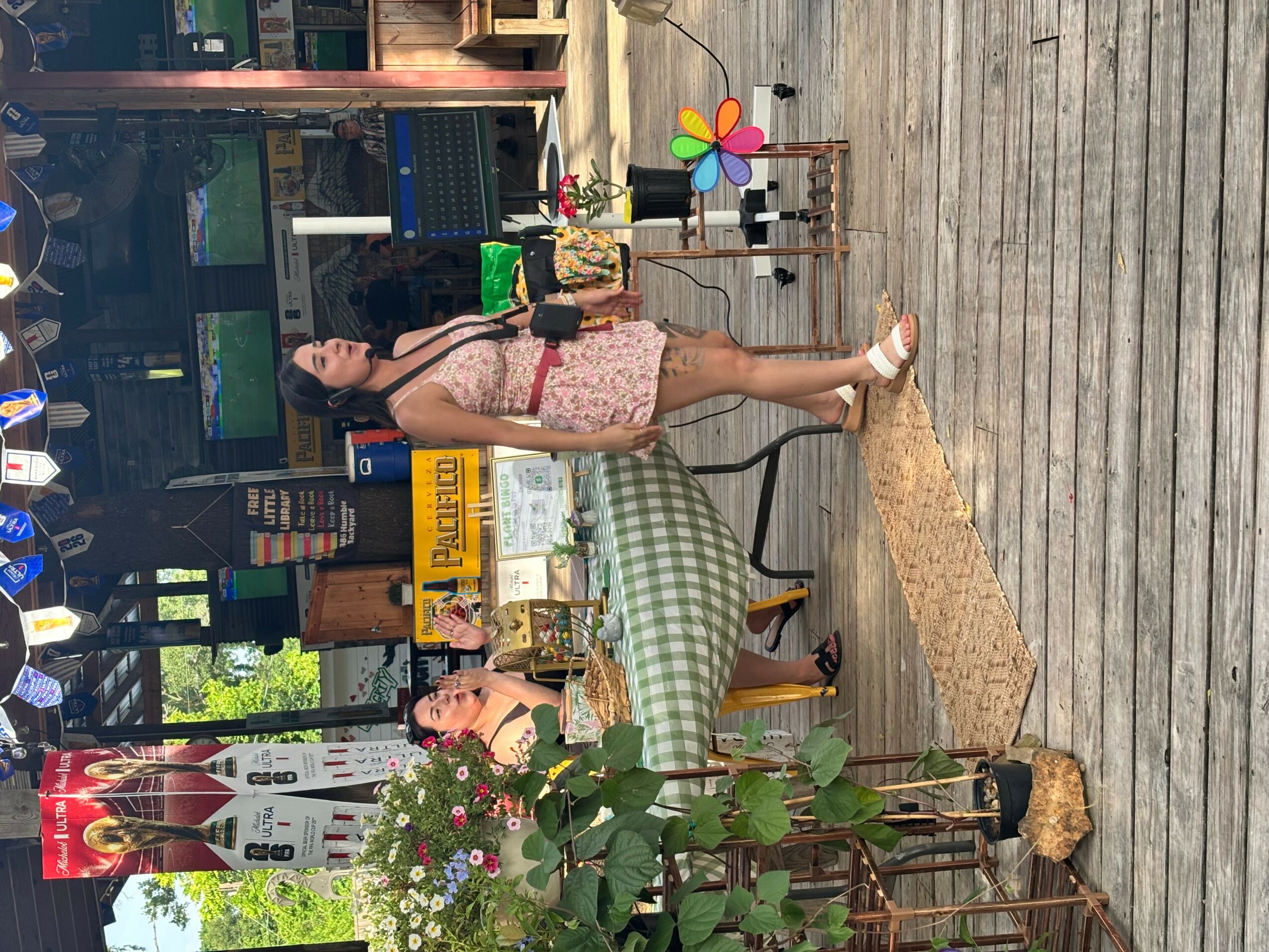 Woman standing on wooden deck near table with plants and decorations, in front of a building with glass windows.