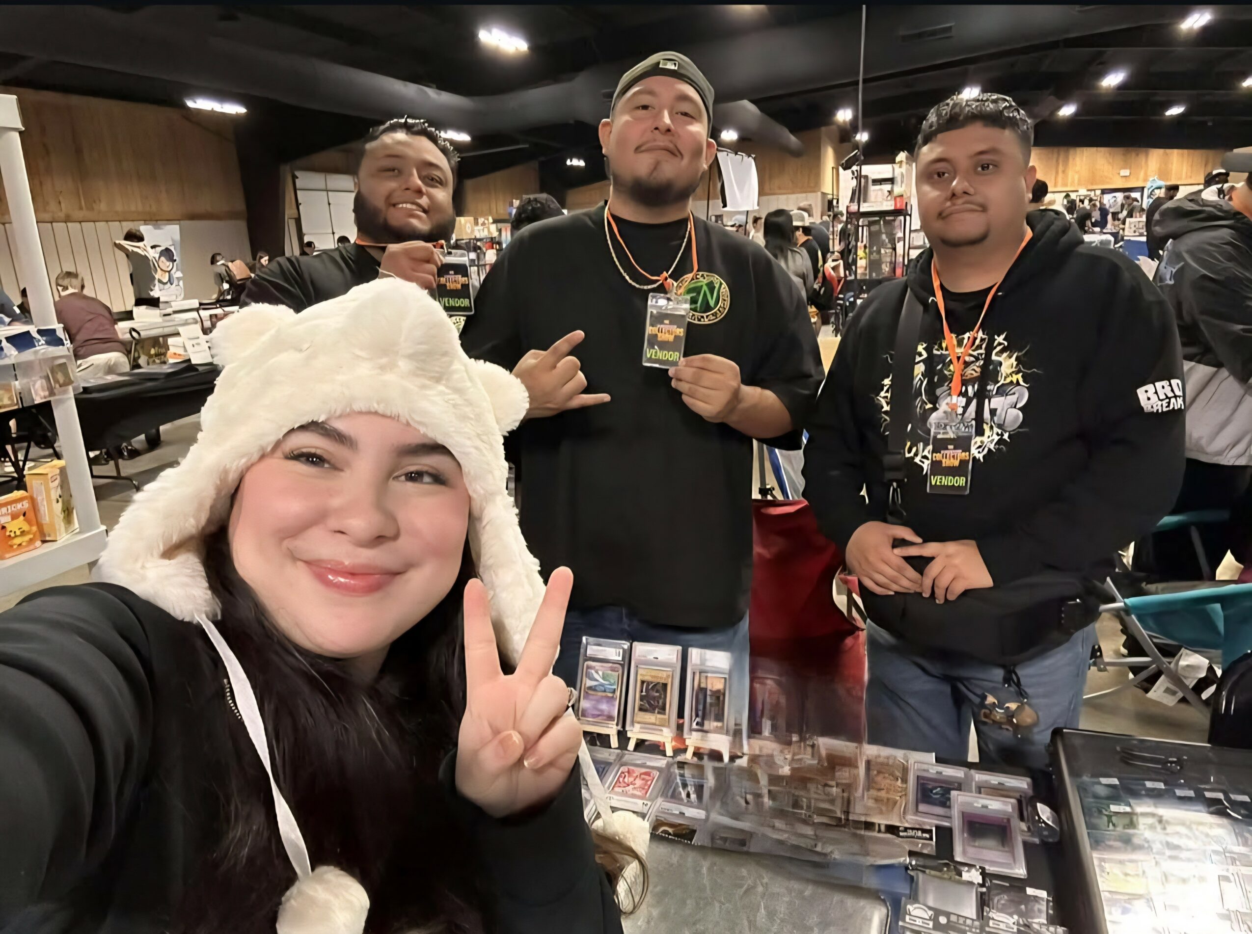 Four people at a convention or event, with one woman in the foreground making a peace sign, wearing a white hat, and three men in the background, one holding a card, in a busy indoor setting.