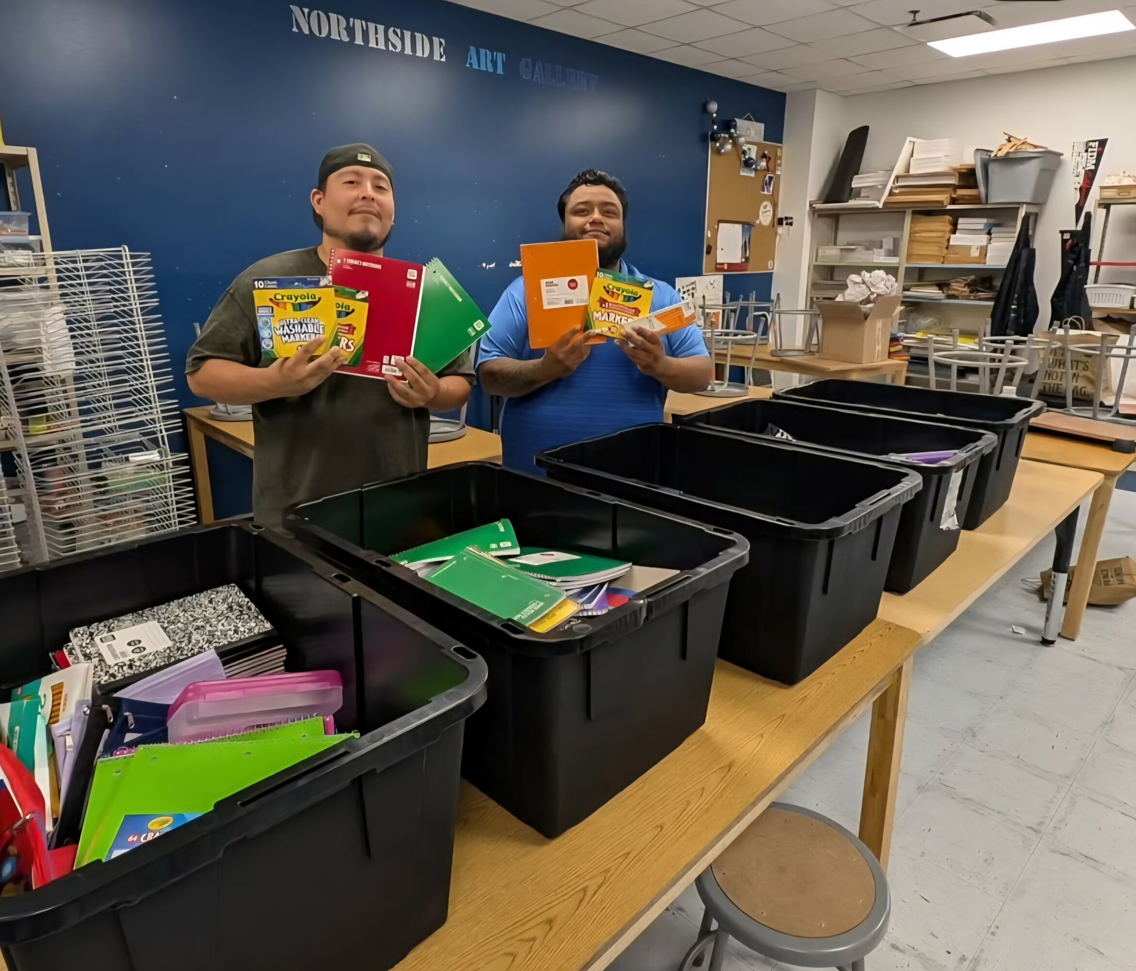 Two people stand behind a table with black bins filled with school supplies in a classroom or office setting.