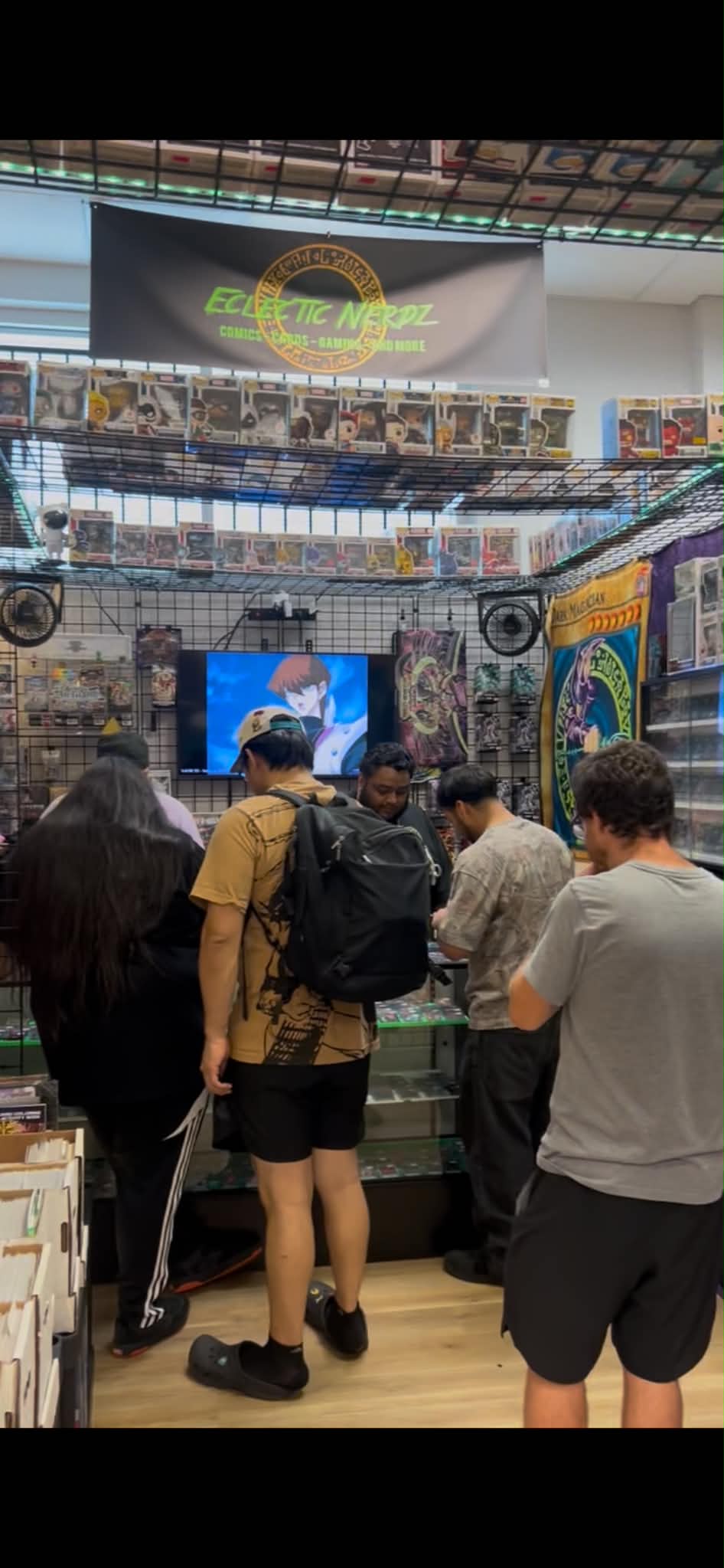 People browsing at a store with shelves and a large screen, under a ceiling with a banner. Four people are visible.