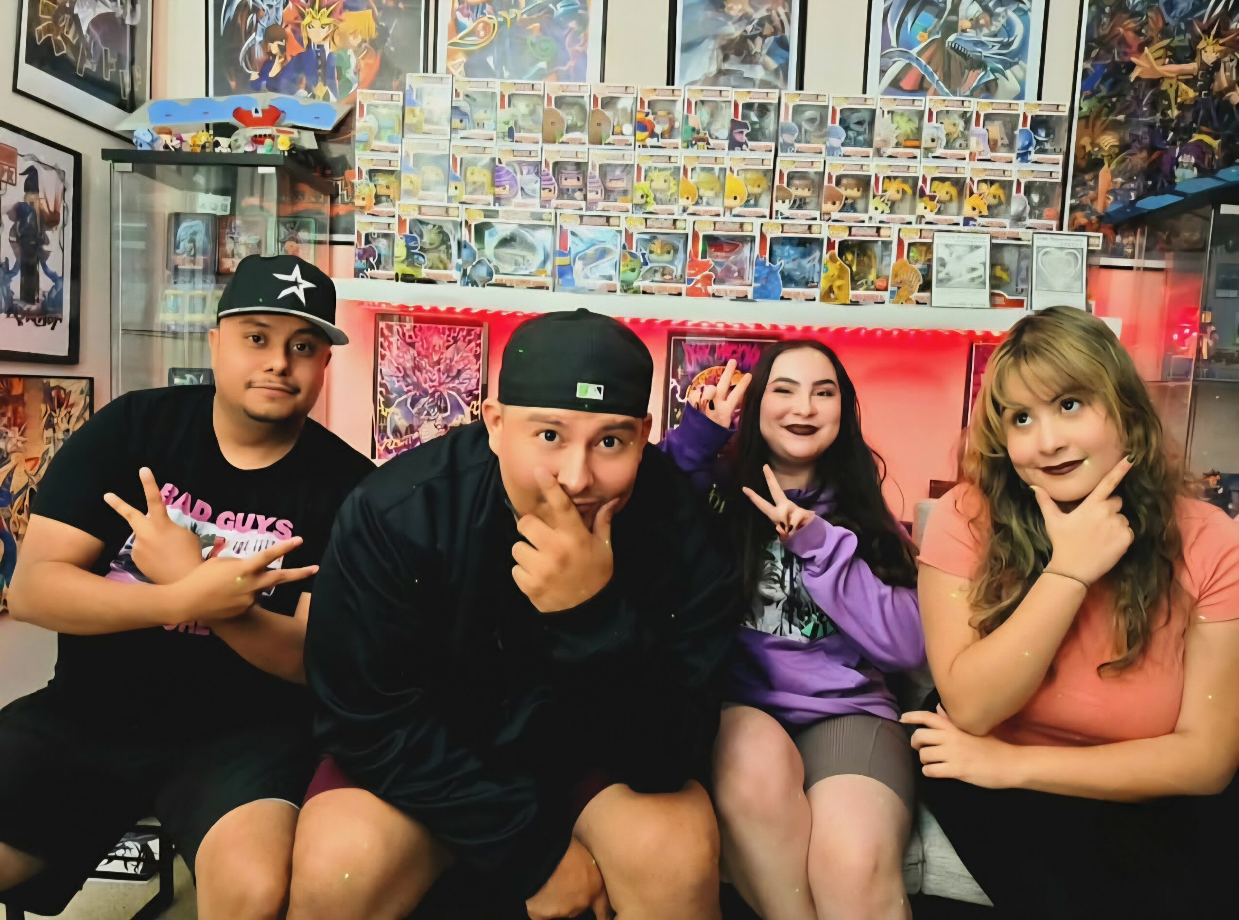 Four people sitting in a store with comic books and posters in the background, smiling and making peace signs.