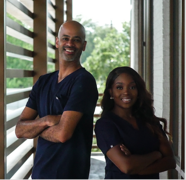 Two healthcare professionals, a man and a woman, standing side by side with arms crossed, smiling, in a bright room.