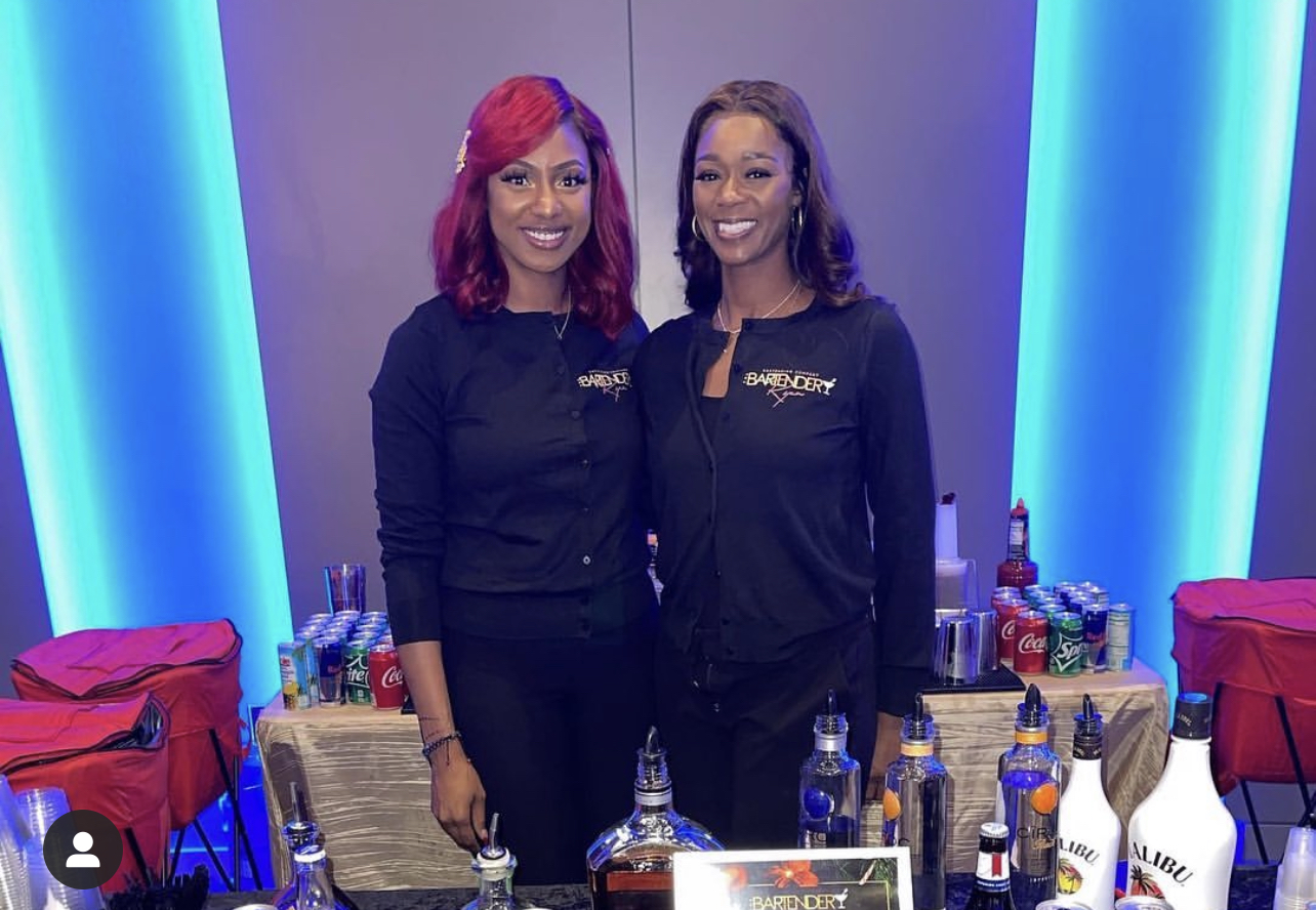 Two women stand behind a table with drinks and supplies, smiling in front of blue-lit background.