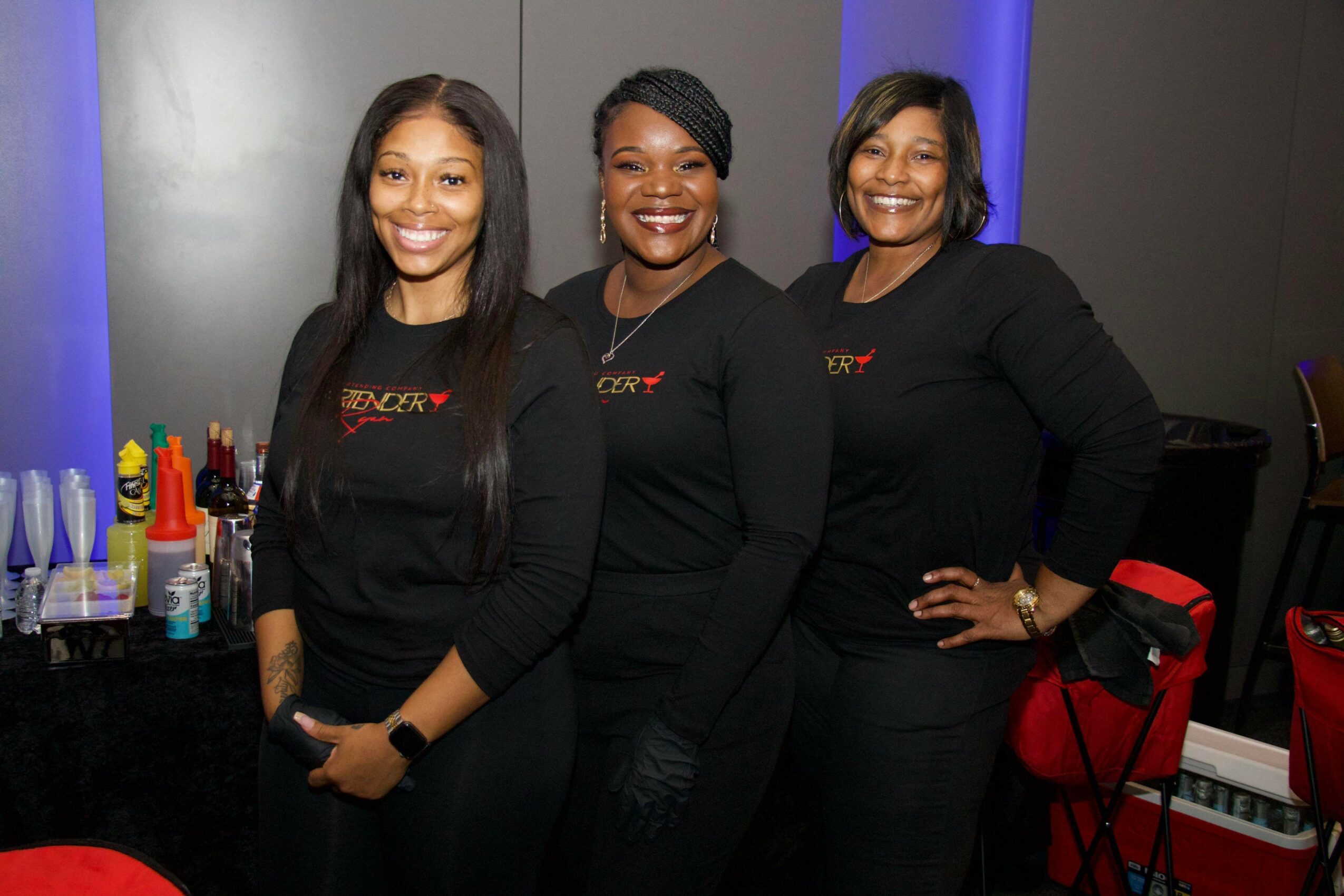 Three women standing together, smiling, in a room with a table of drinks and a dark background.