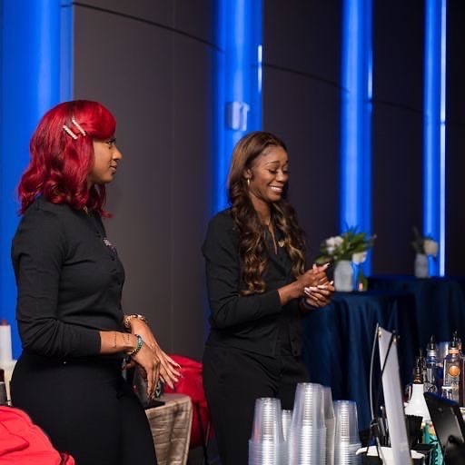 Two women standing indoors, one smiling and holding a small plant, with blue lighting and tables in the background.