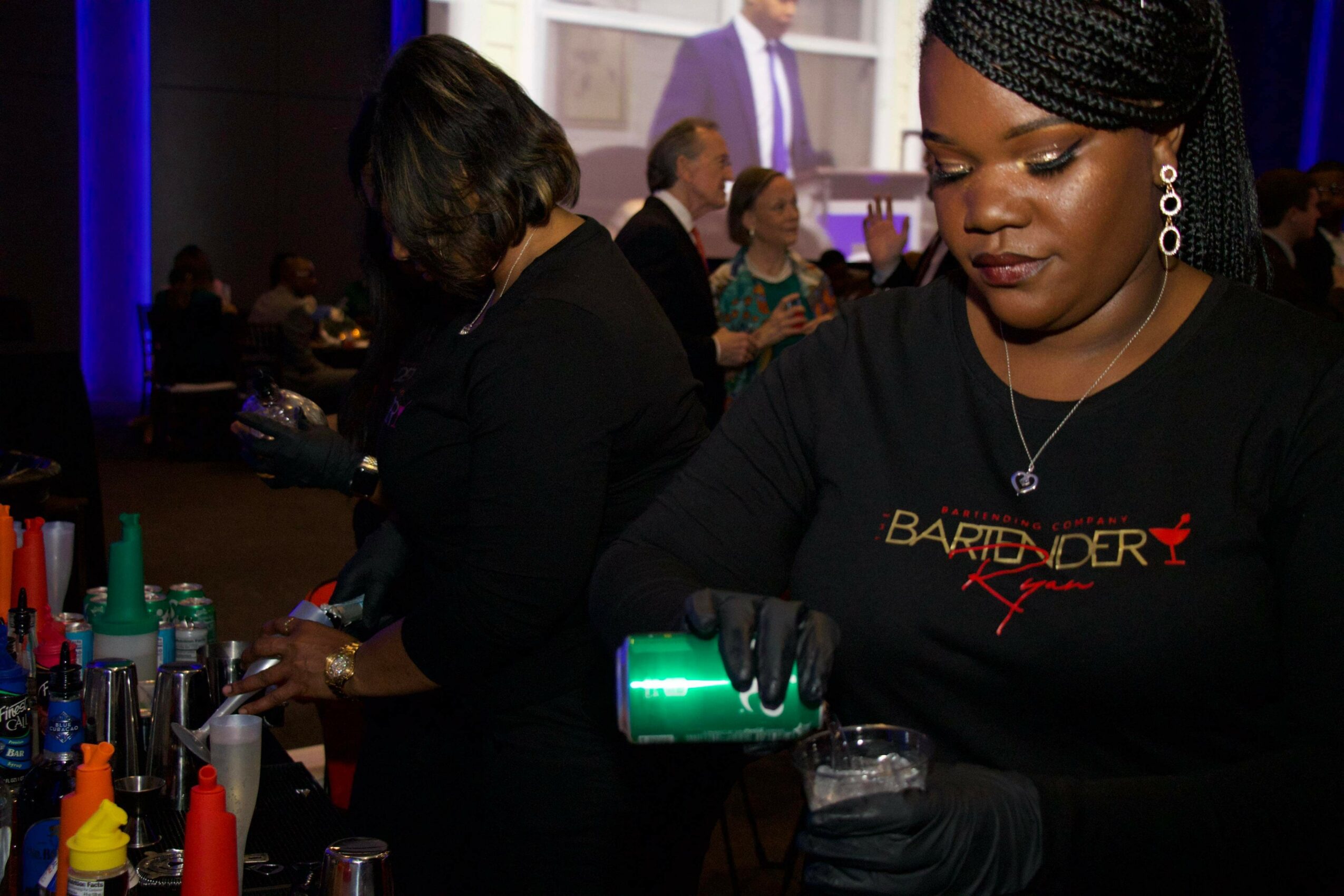 Woman with braided hair pouring a drink at an event, with other people in the background.