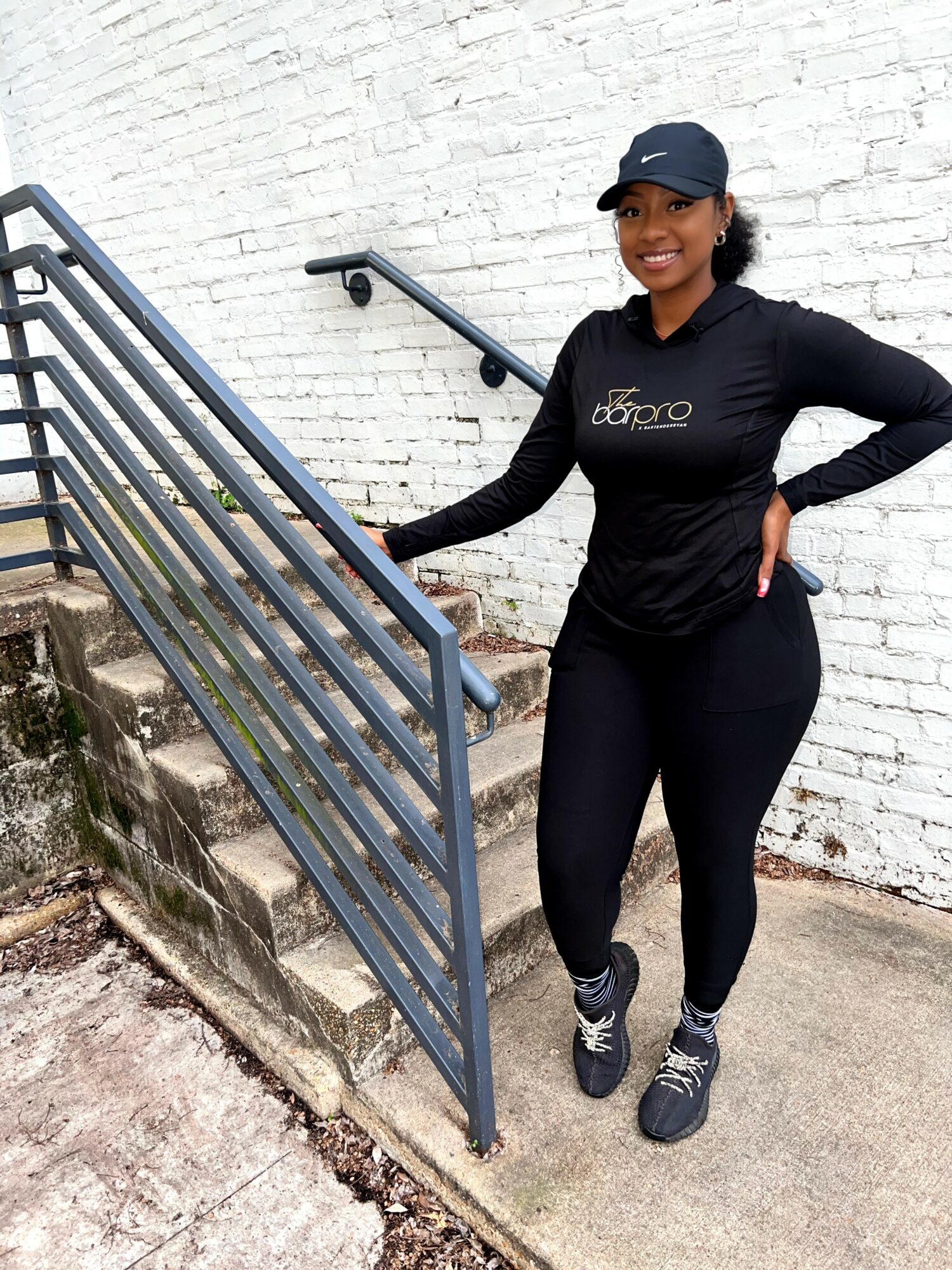 Smiling woman in athletic clothing and cap standing on stairs outside, holding railing, against white brick wall.