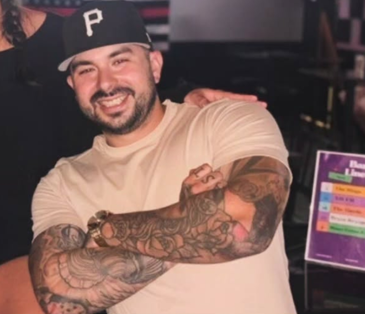 Smiling man with tattoos on arms, wearing a beige shirt and black cap, in a social setting.