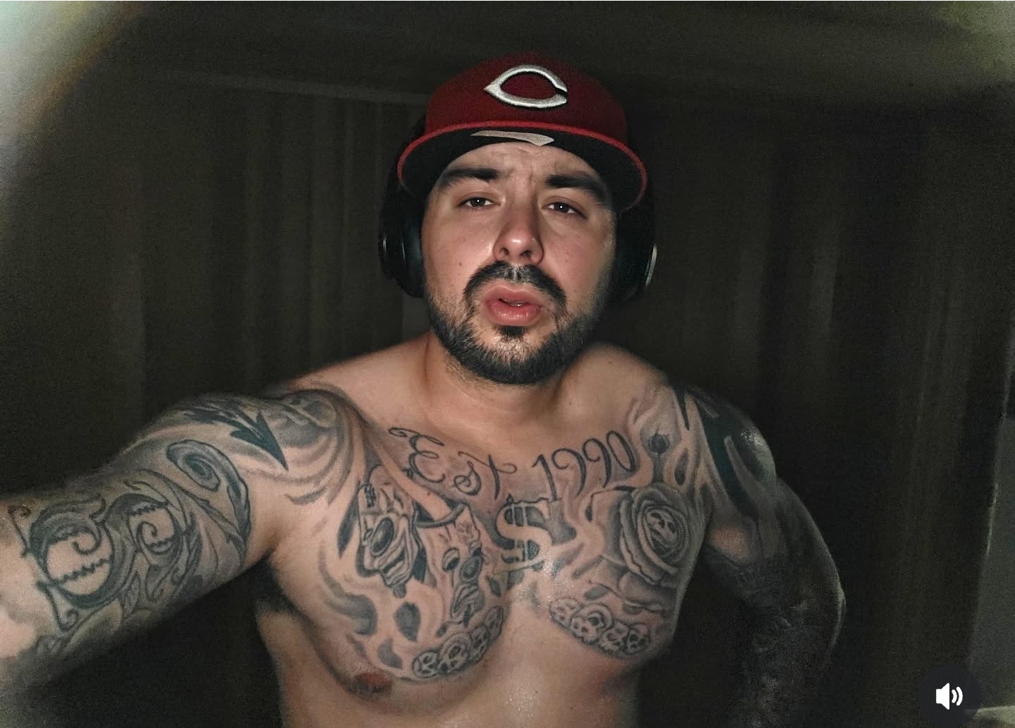 Young man with tattoos on chest and arms, wearing a red baseball cap and headphones, taking a selfie indoors.