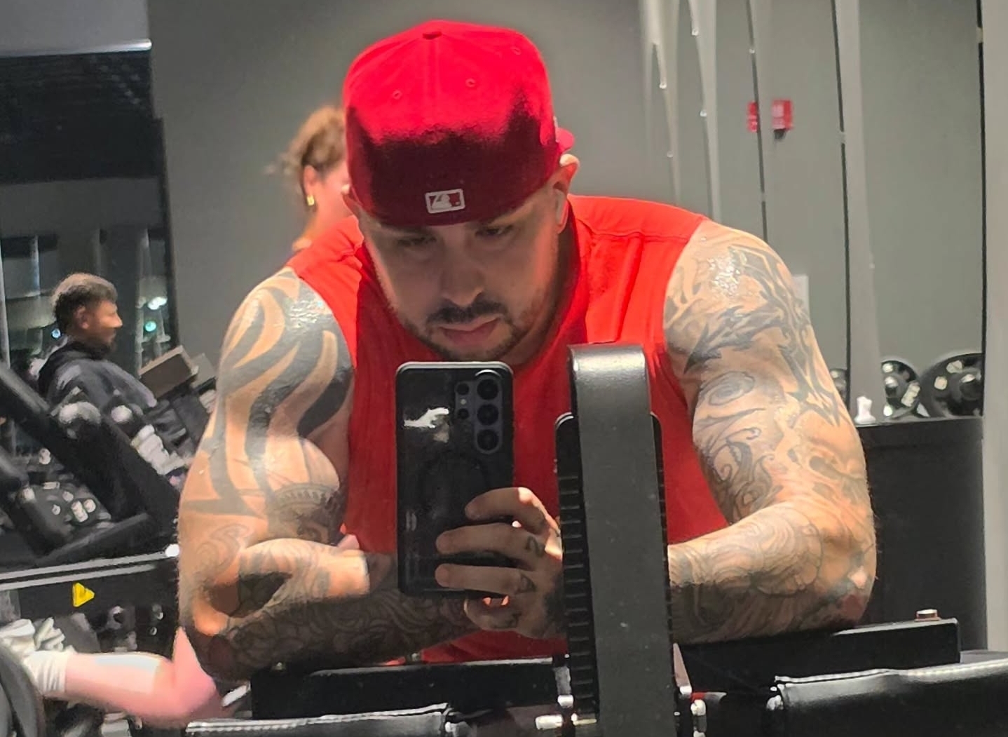Man with tattoos wearing a red cap and sleeveless shirt taking a mirror selfie in gym.
