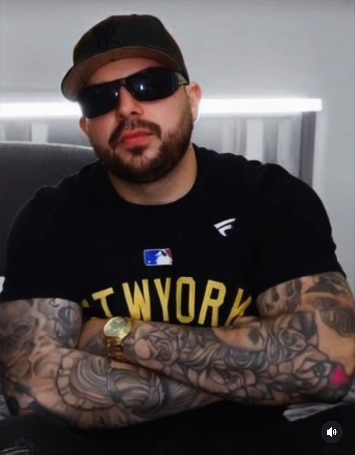 Man with tattoos, sunglasses, and a cap, crossing arms, wearing a black T-shirt with yellow text, in an indoor setting.