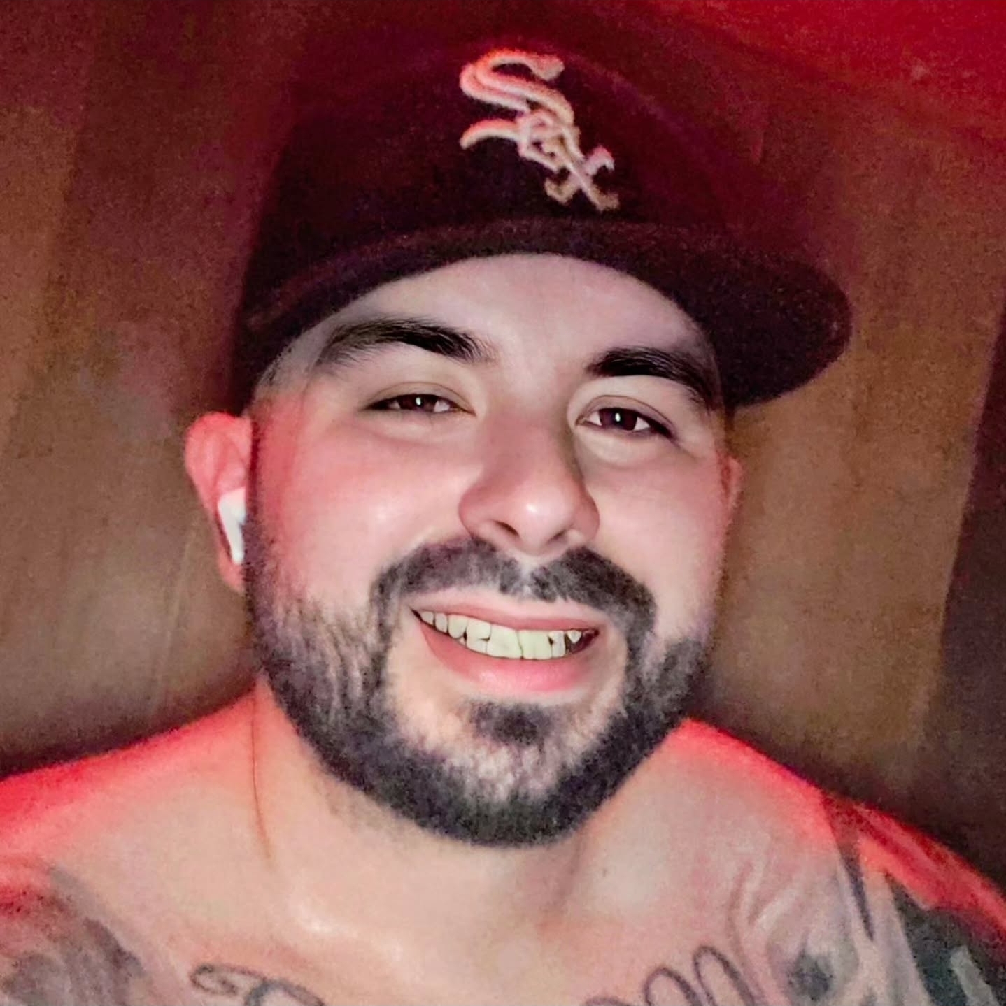 Smiling man with a beard wearing a black baseball cap with a white logo, earbud, and tattoos on his chest.