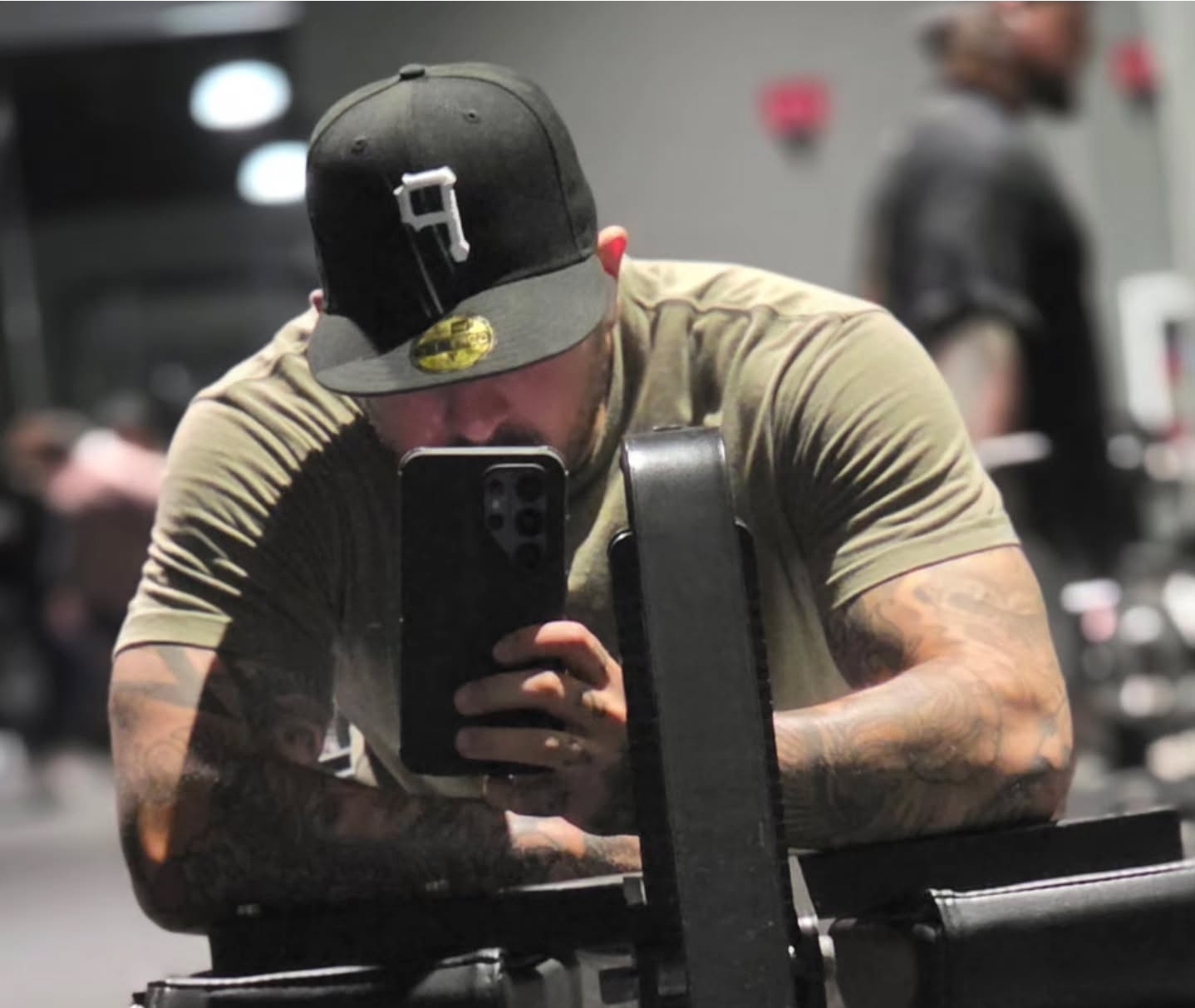 Person taking a mirror selfie at a gym, wearing a black cap and a beige shirt, with a backpack on shoulder.