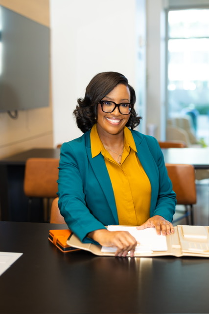 Woman with glasses and curly hair smiling, sitting at a desk with open book, in a bright office space.