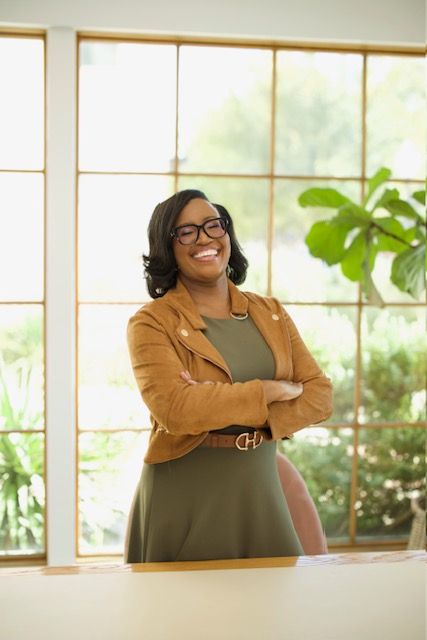 Woman with glasses and dark hair, smiling, standing near a window with green plants outside.