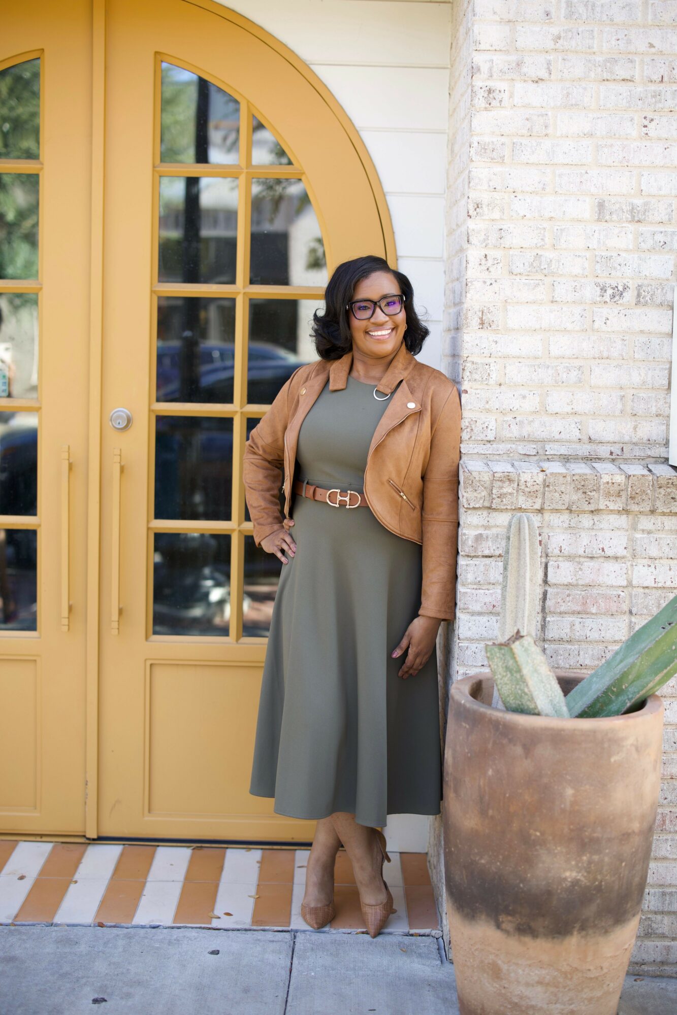 Woman standing outside near a yellow door, smiling, wearing glasses, a brown jacket, and a long gray skirt.