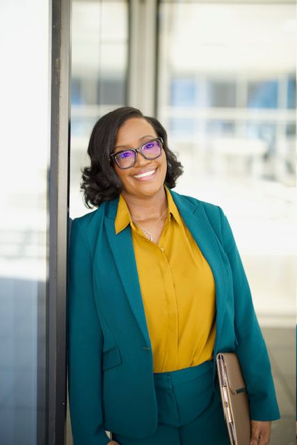 Woman with glasses smiling, wearing a teal blazer and yellow blouse, standing in front of a window with city buildings behind her.