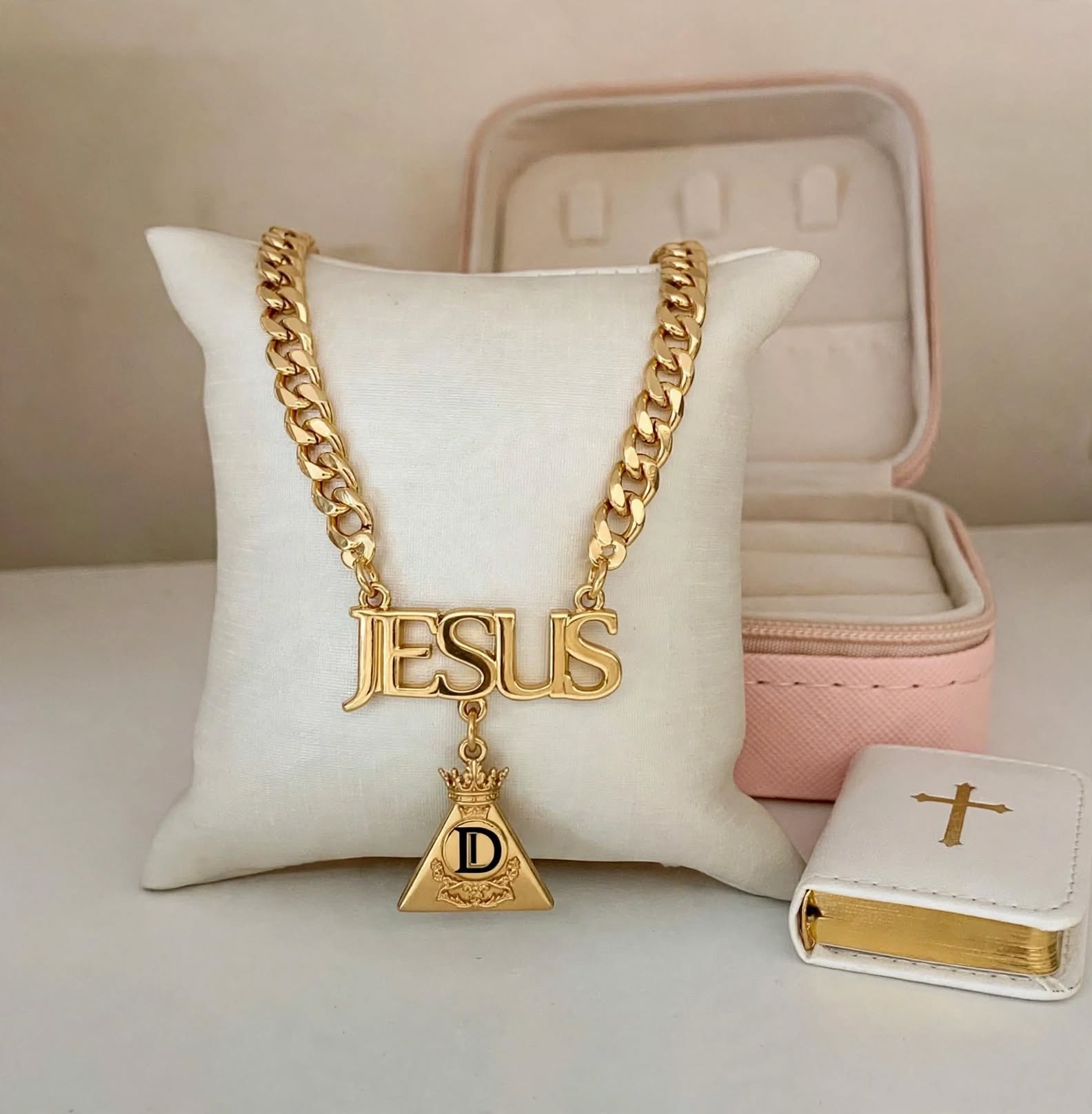 Gold chain necklace with 'JESUS' pendant and a small book with a cross, displayed on a white pillow.
