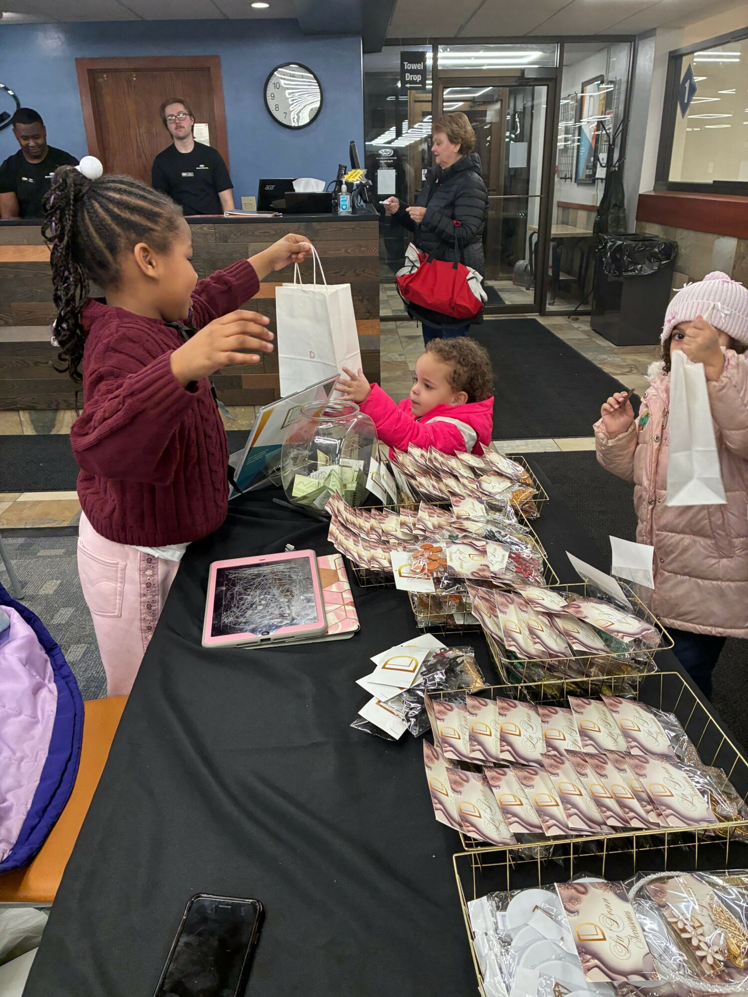 Children and adults around a table with packaged baked goods, in a room with glass doors and a clock on the wall.