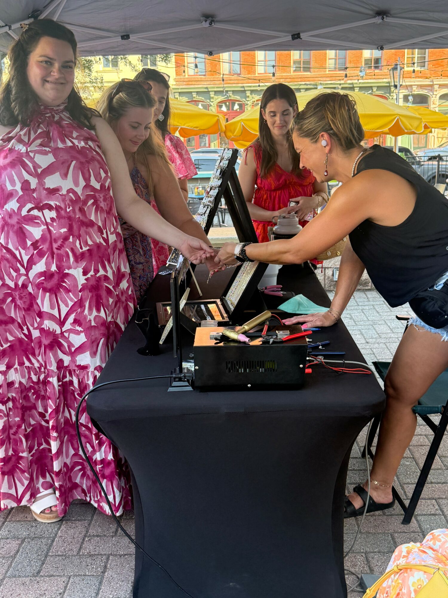 Group of women at a booth with electronic devices, some smiling, outdoors with yellow umbrellas and brick buildings in background.