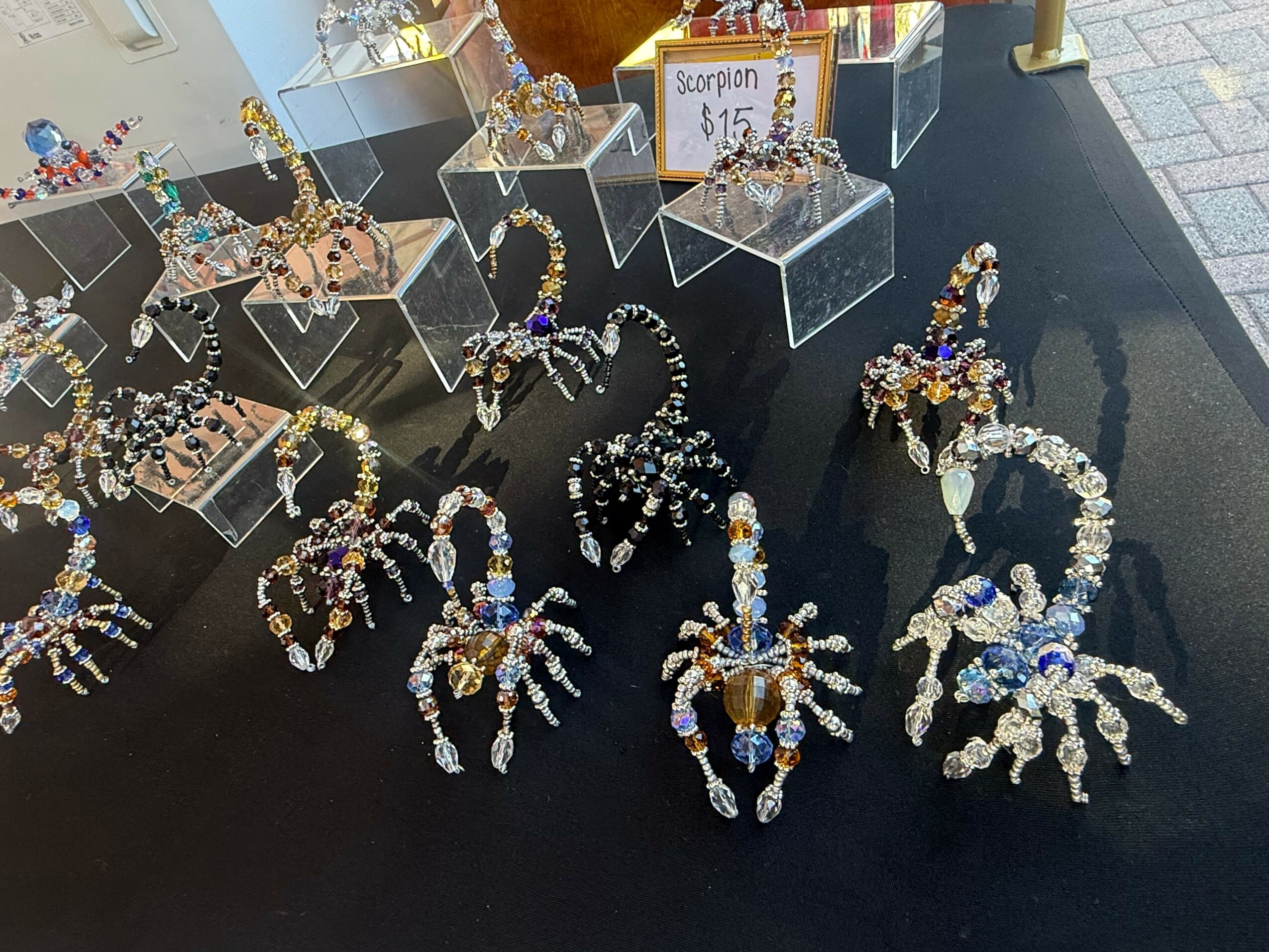 Display of jewelry including bracelets and necklaces on a black table with clear acrylic stands, and a sign showing a scorpion for $15.