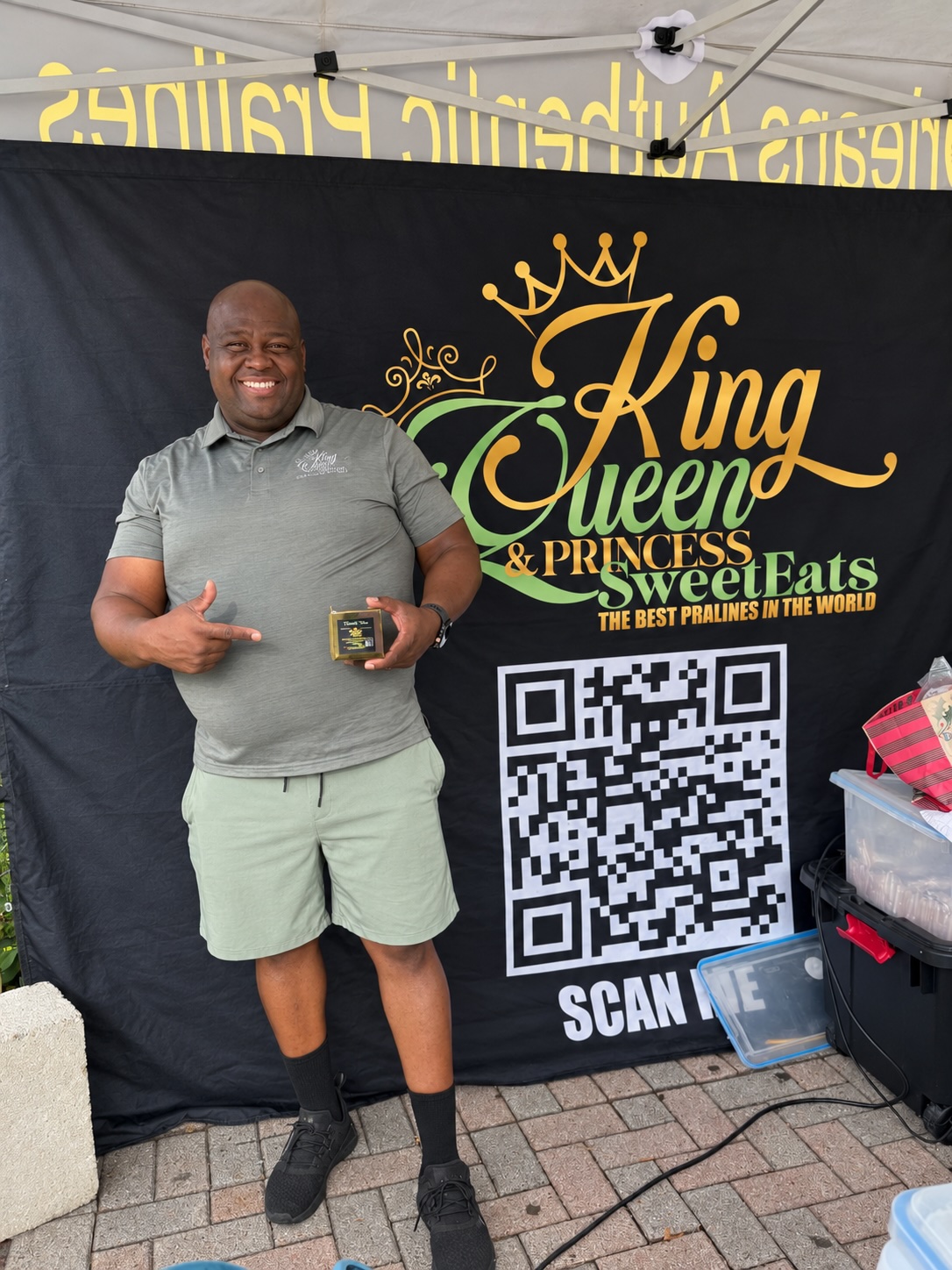 Smiling man in gray shirt and shorts holding a small container, standing in front of a black backdrop with colorful text and a QR code.