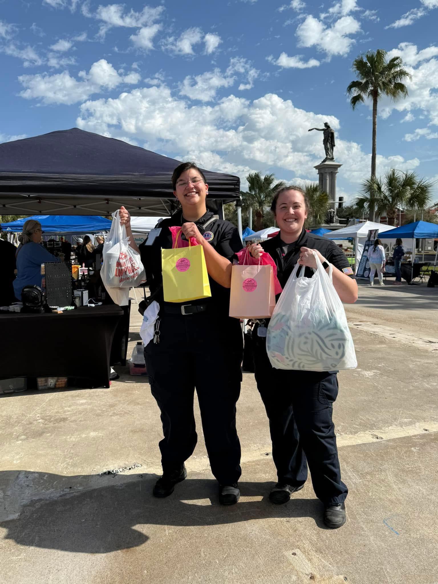 Two women stand outdoors holding shopping bags, with tents, a statue, and palm trees in the background under a partly cloudy sky.