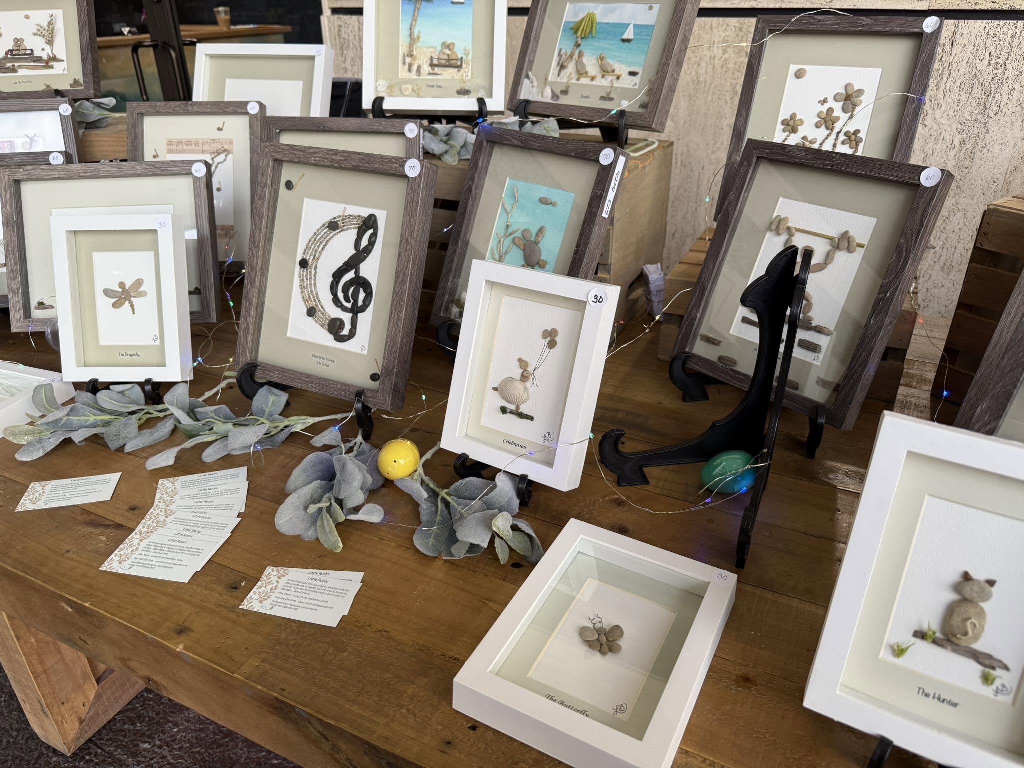 Display of framed artwork on a wooden table, with decorative items and notes, in a gallery setting.