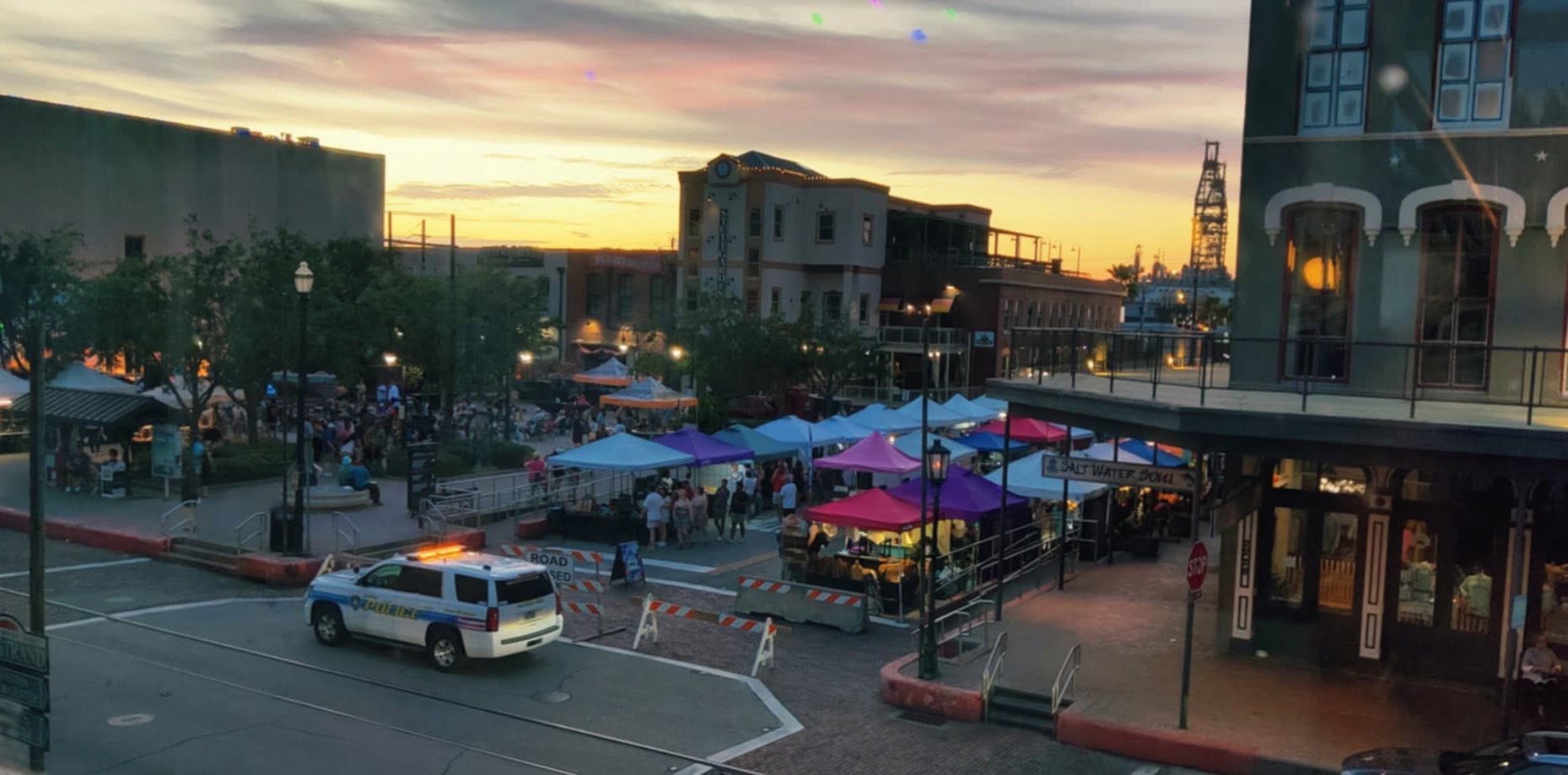 Outdoor market with tents and people, city buildings, and a sunset sky, viewed from across the street.
