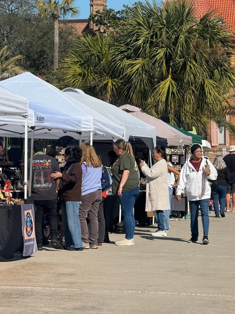People browsing outdoor market stalls with white tents, trees, and buildings in the background.