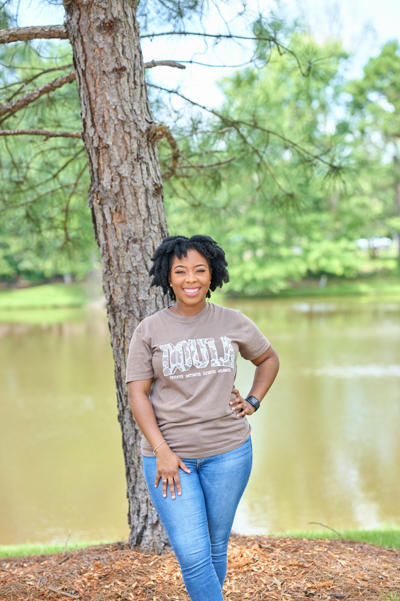 Smiling woman with curly hair standing outdoors near a tree by a lake, wearing a gray t-shirt and blue jeans.
