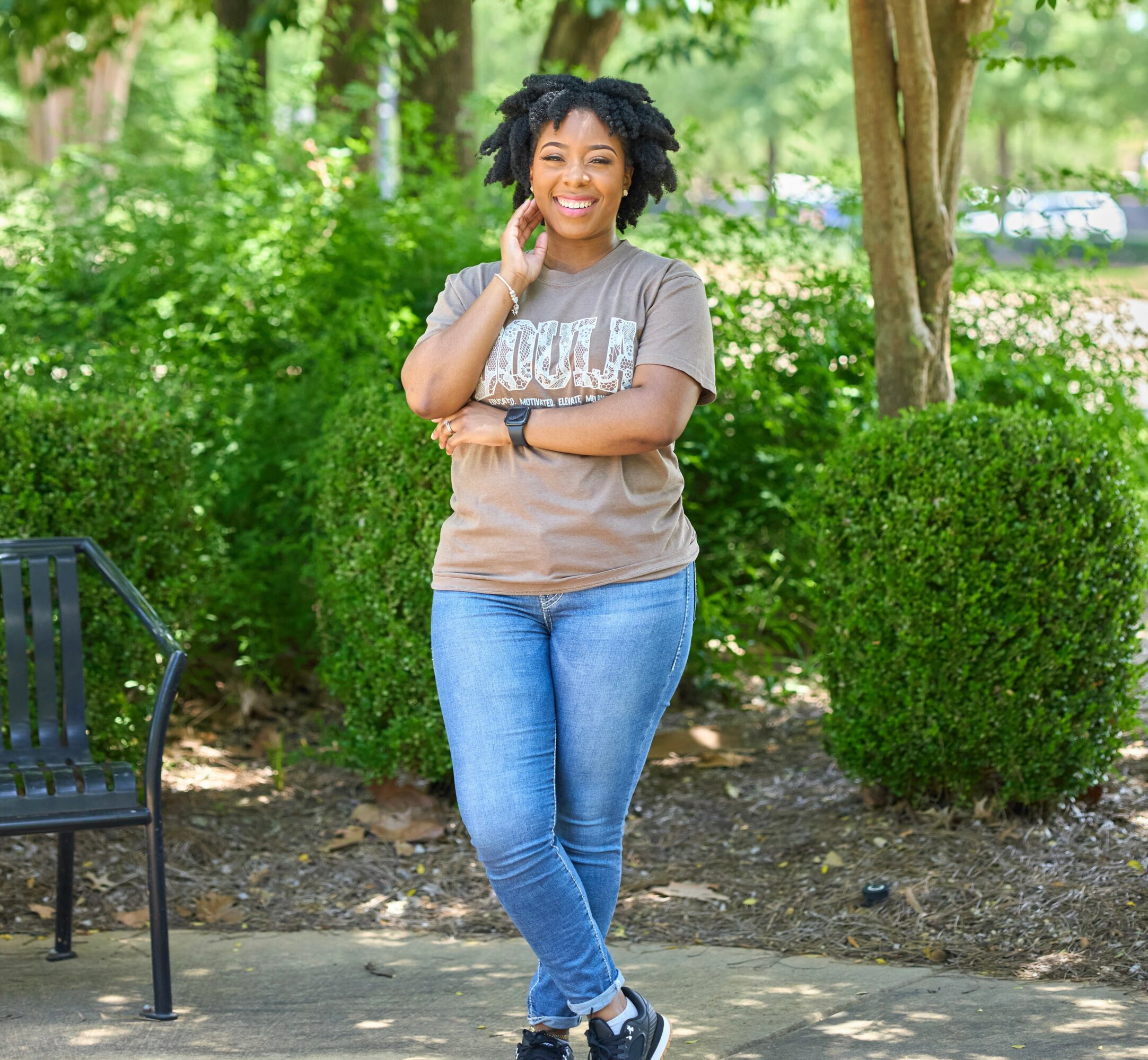 Young woman with curly hair smiling outdoors, standing with arms crossed, surrounded by greenery and trees.