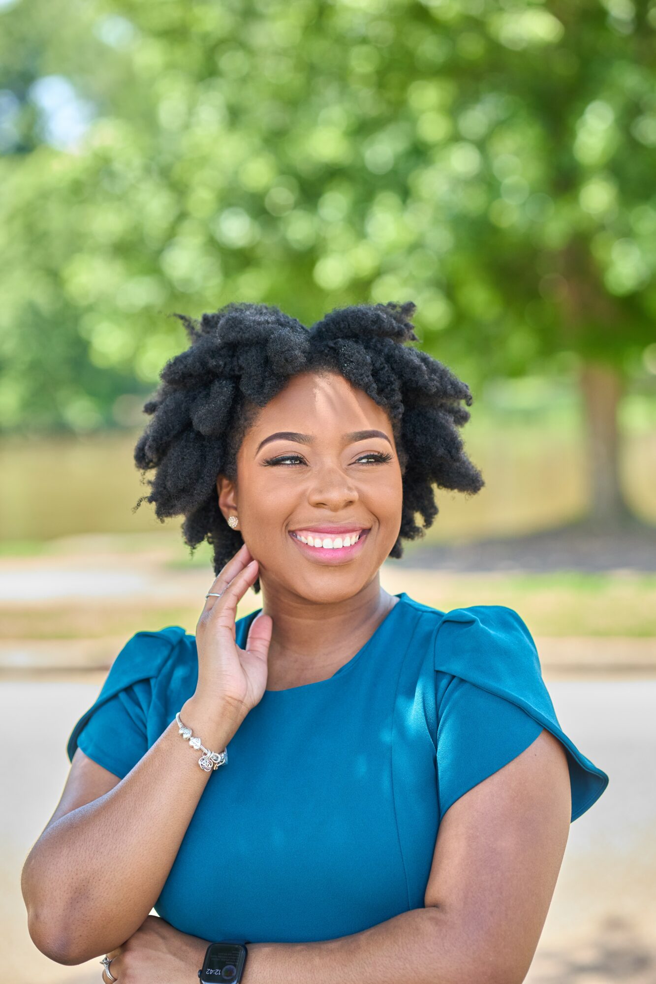 Smiling woman with natural hair in a blue dress outdoors, green trees in background.