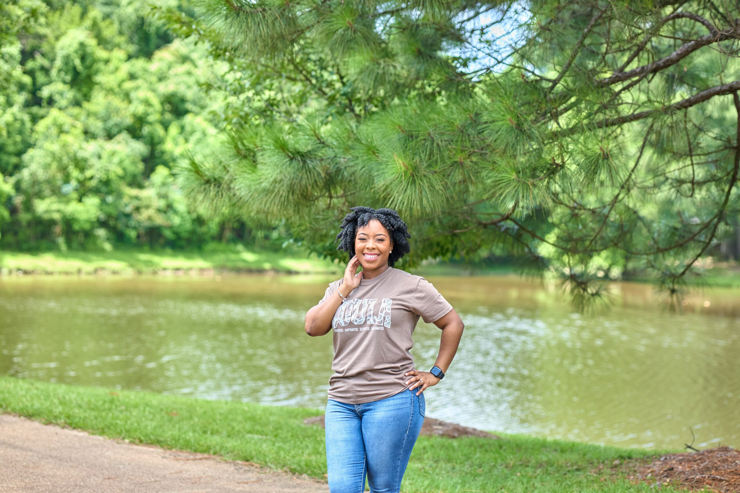 Woman standing near a lake with trees in the background, smiling, wearing a gray t-shirt and blue jeans.