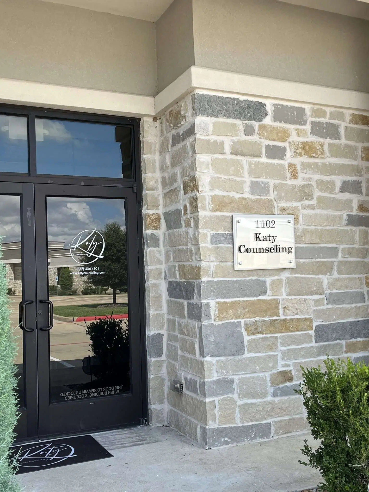 Entrance door with a sign reading 'Katy Counseling' on a brick wall, with a small bush nearby.