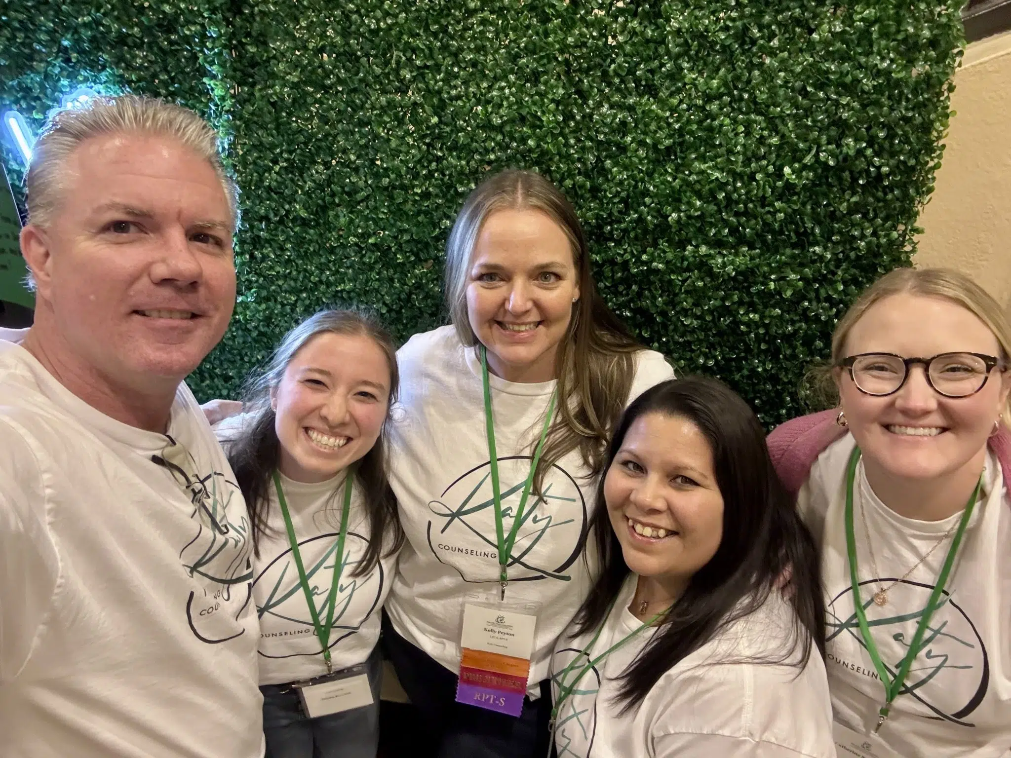 Group of five smiling people wearing white shirts with green lanyards, standing in front of a green hedge wall.
