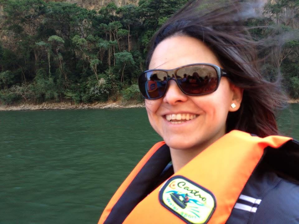 Smiling woman wearing sunglasses and life jacket outdoors near water with trees in background.