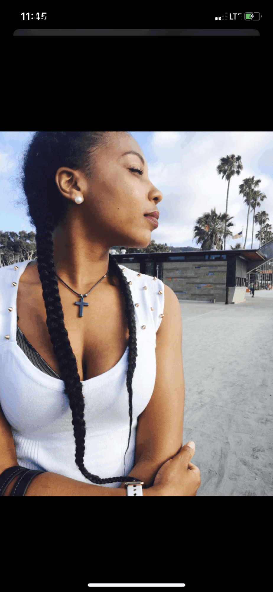 Young woman with braided hair wearing a white top and cross necklace, standing outdoors with palm trees in background.