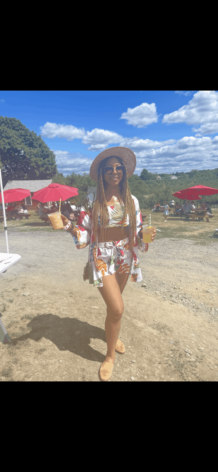 Person in a floral outfit and hat holding drinks outdoors with umbrellas and trees, blue sky, and clouds in background.