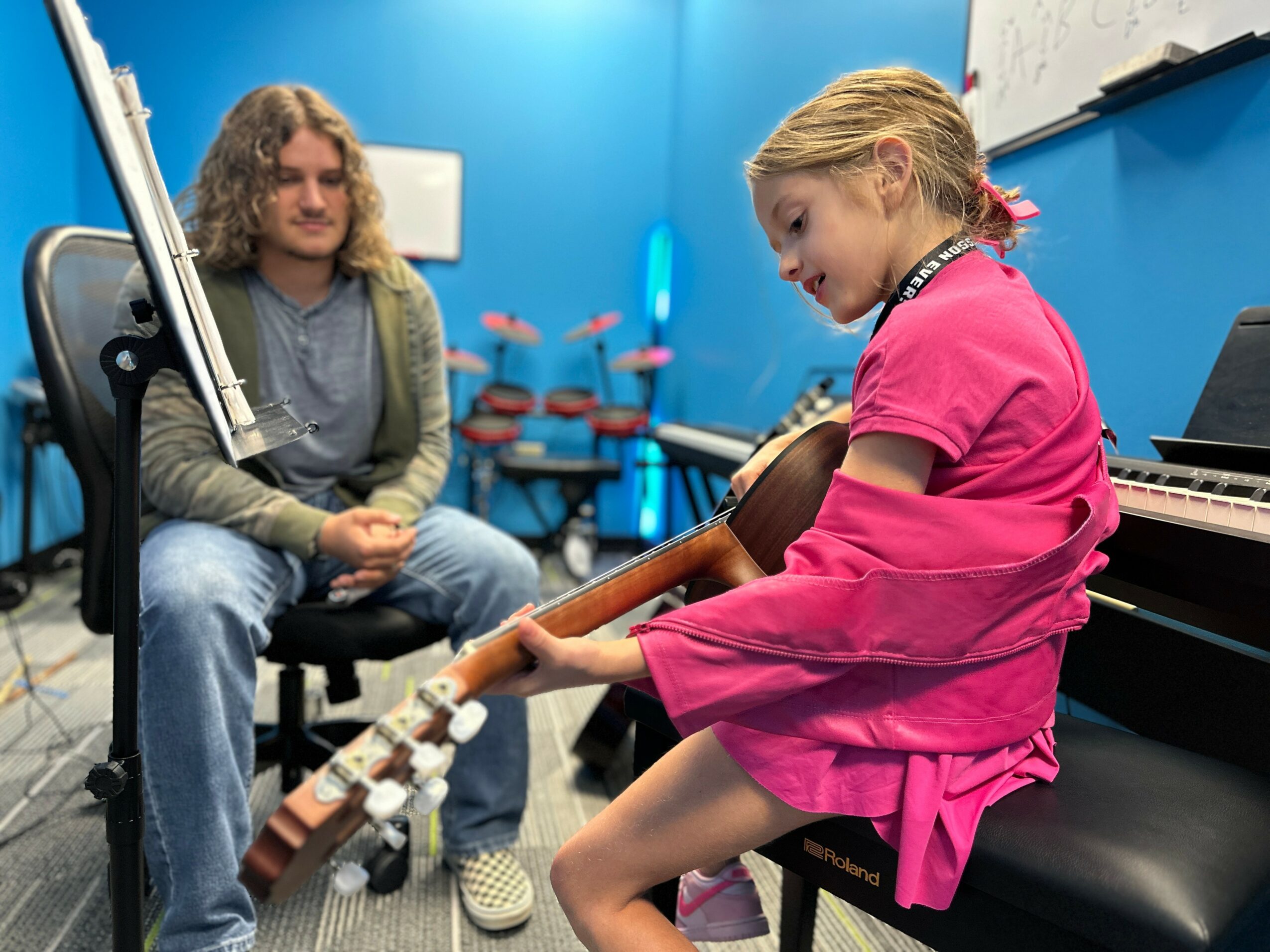 Young girl in pink dress holding a guitar, sitting on a bench, with a man in a music room. 20 words.