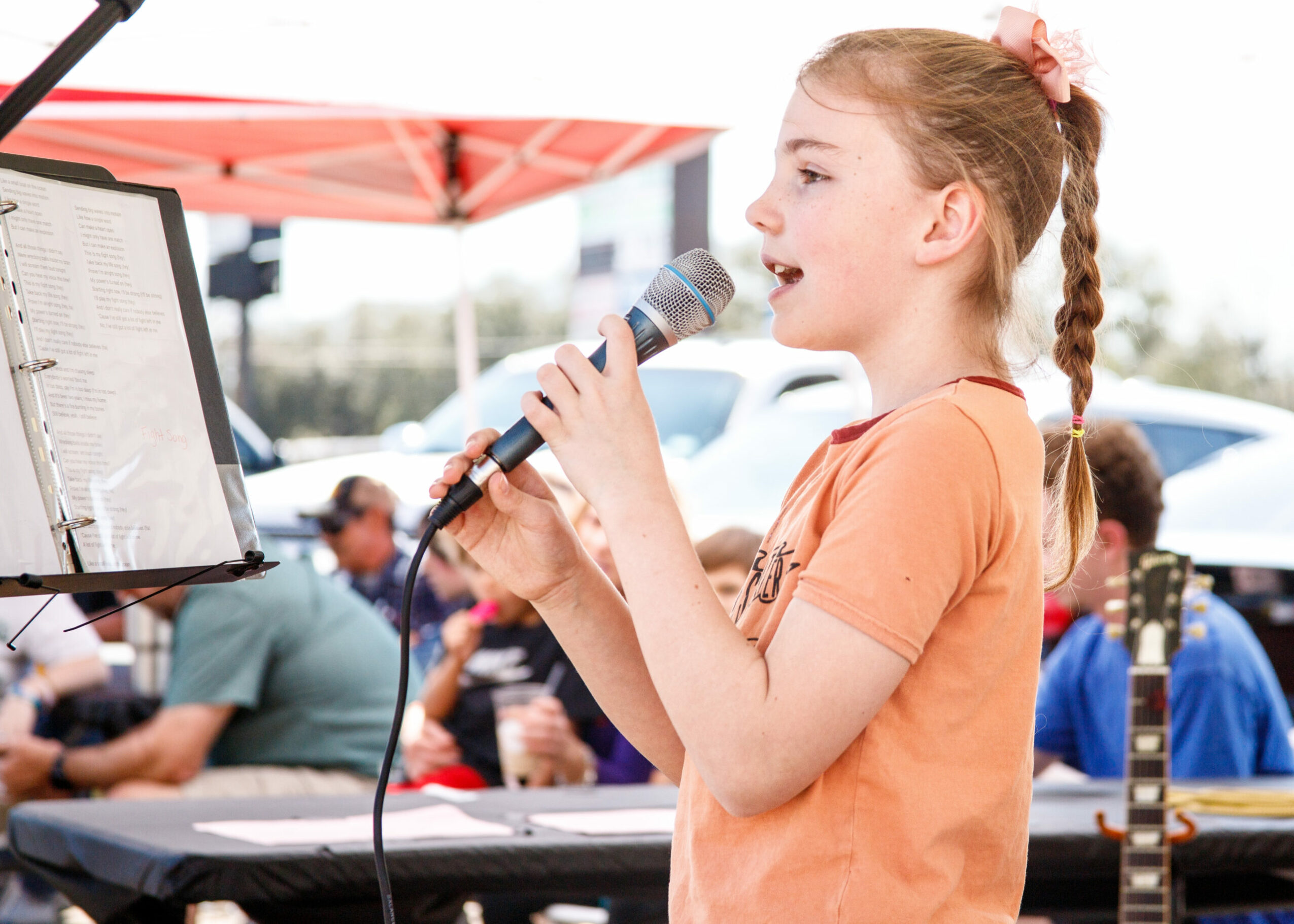 Young girl singing into a microphone outdoors with a music stand and people in the background.