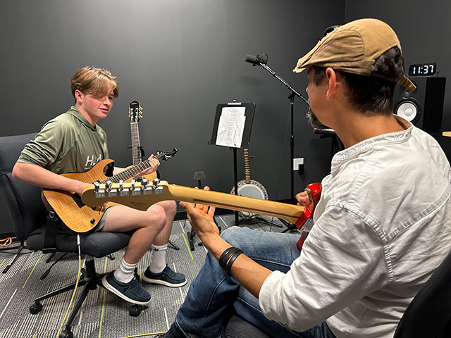 Two young men playing guitars in a music studio with a music stand and microphone, one facing the camera, the other with back to it.