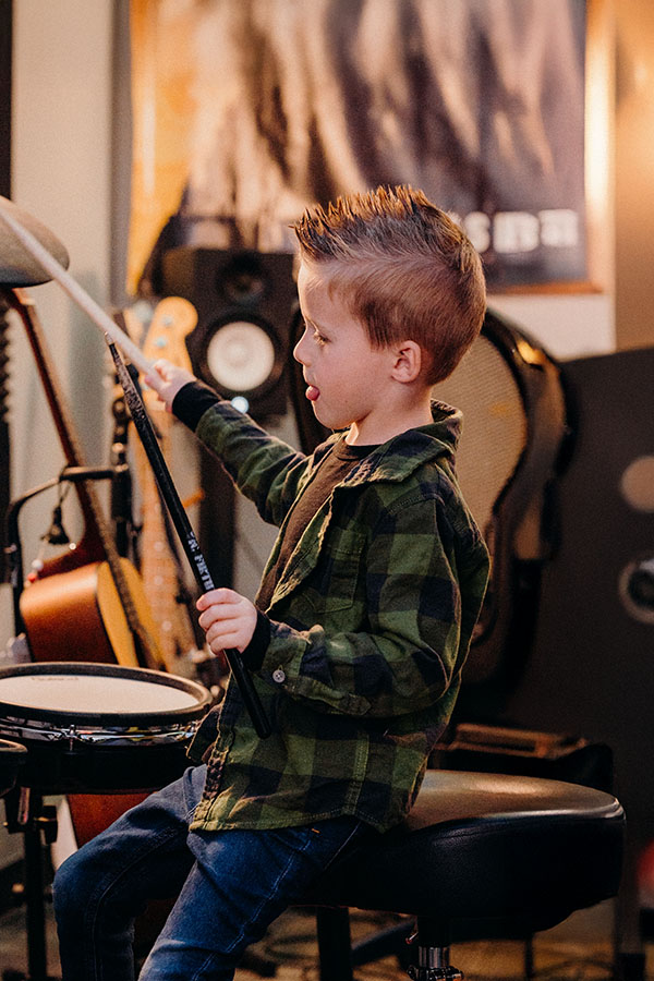 Child with spiked hair playing drumsticks on a drum in a music room, surrounded by speakers and musical equipment.