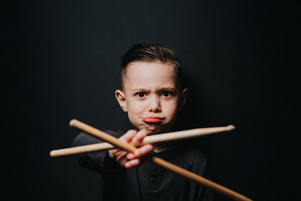 Boy with a serious expression holding two drumsticks crossed in front of him against a dark background.