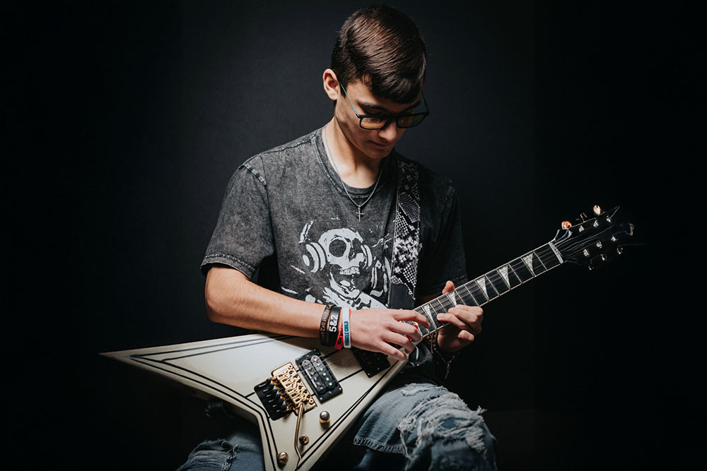 Young man with glasses playing an electric guitar against a dark background.