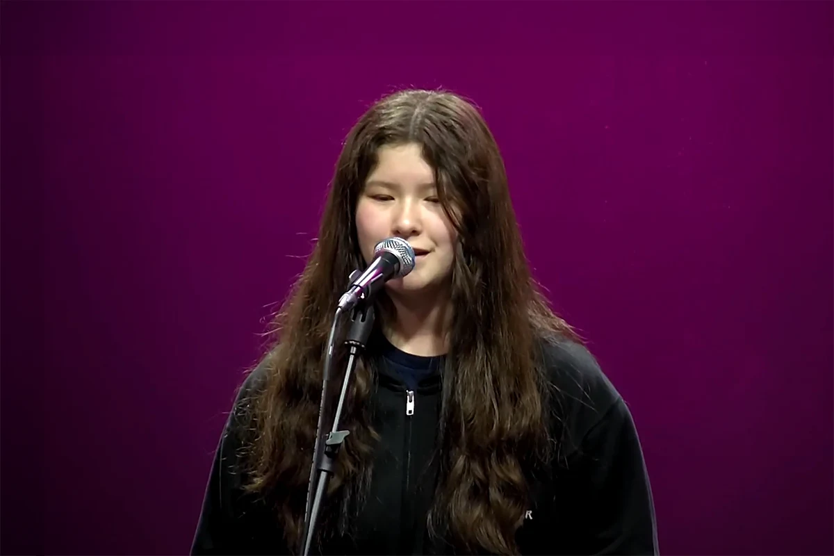 Young girl with long hair speaking into a microphone on stage with a purple background.