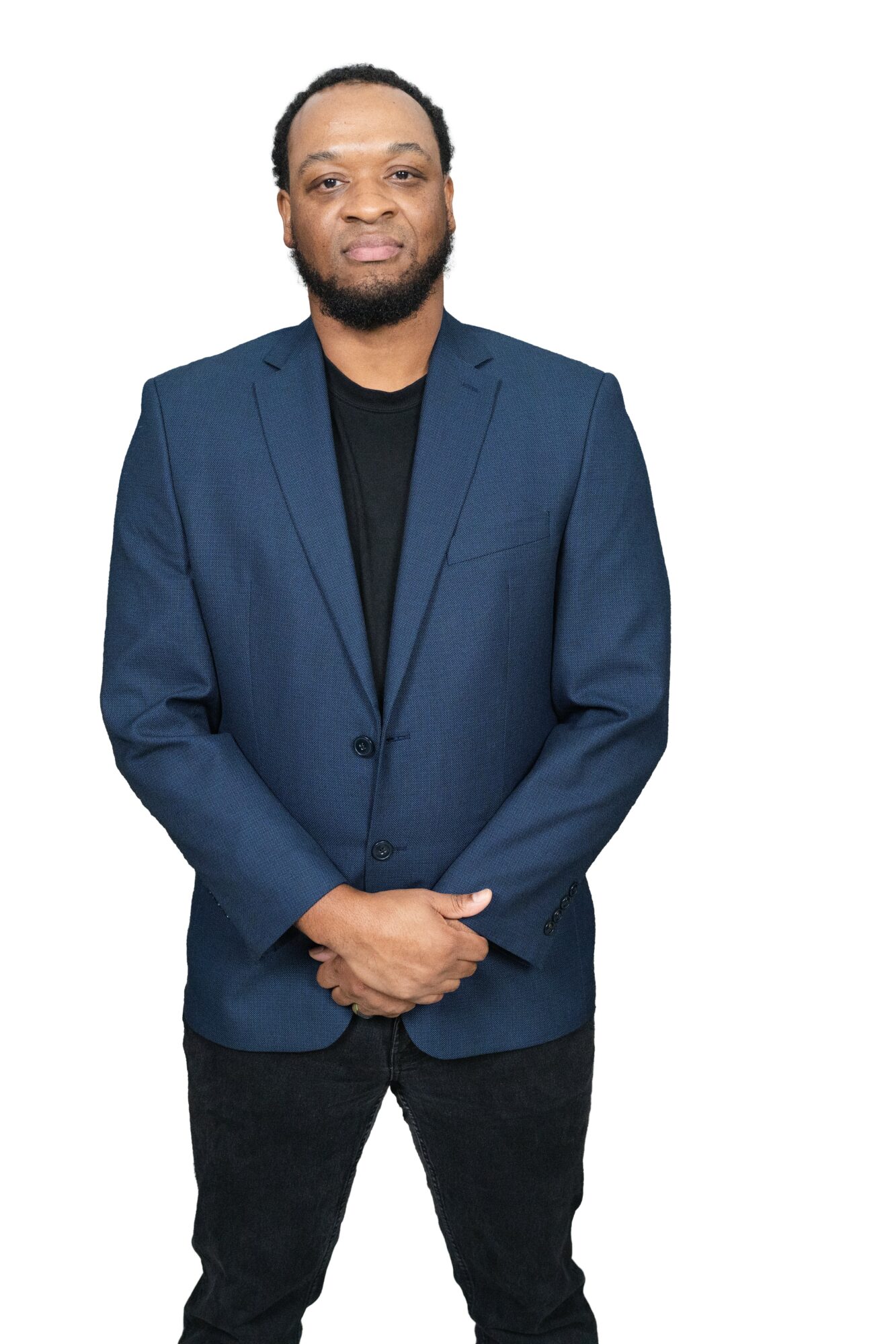 Man with a beard in a blue blazer and black shirt, standing with hands clasped, smiling, against a white background.